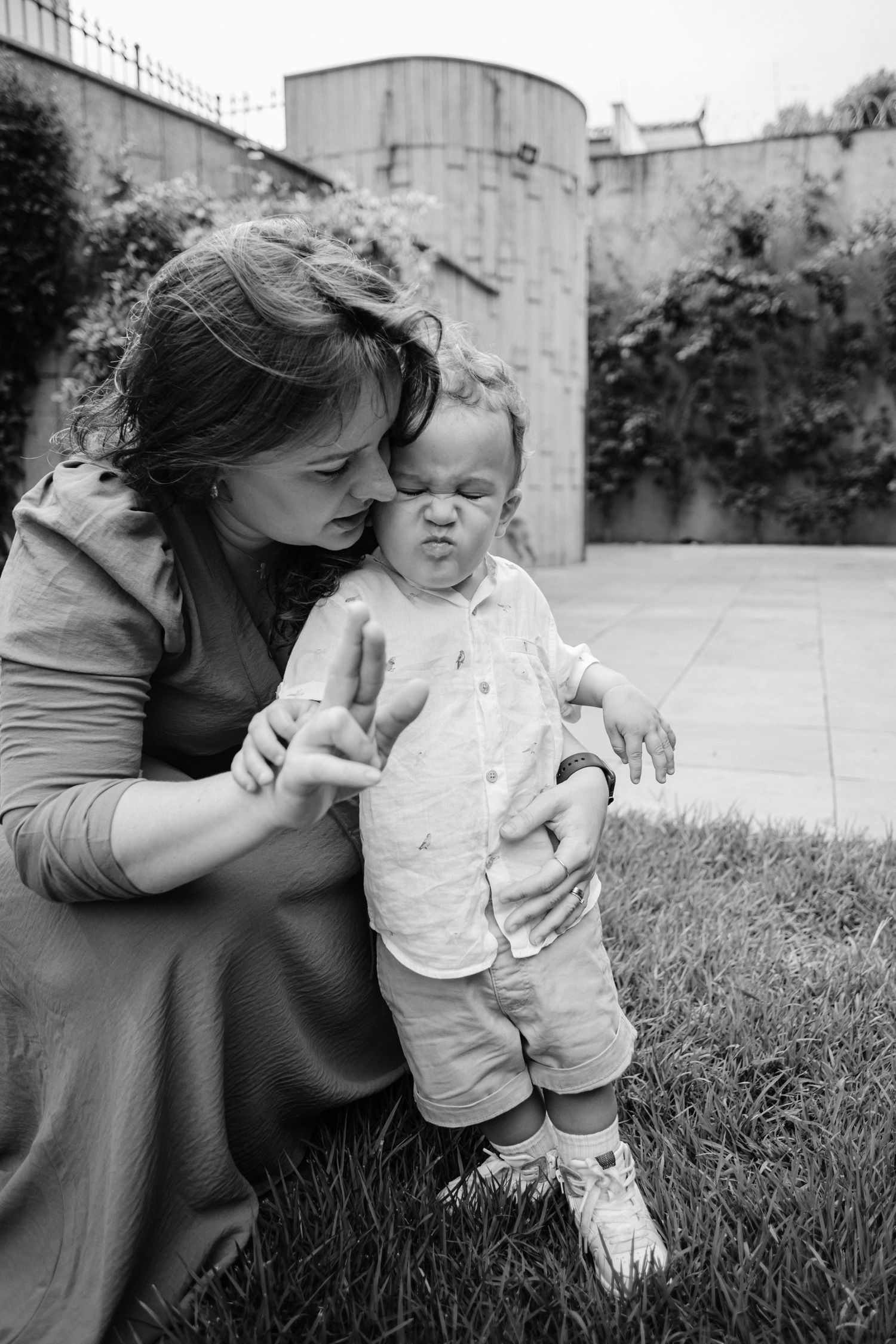 Black and white candid portrait of parent and toddler interacting outdoors by an industrial backdrop.