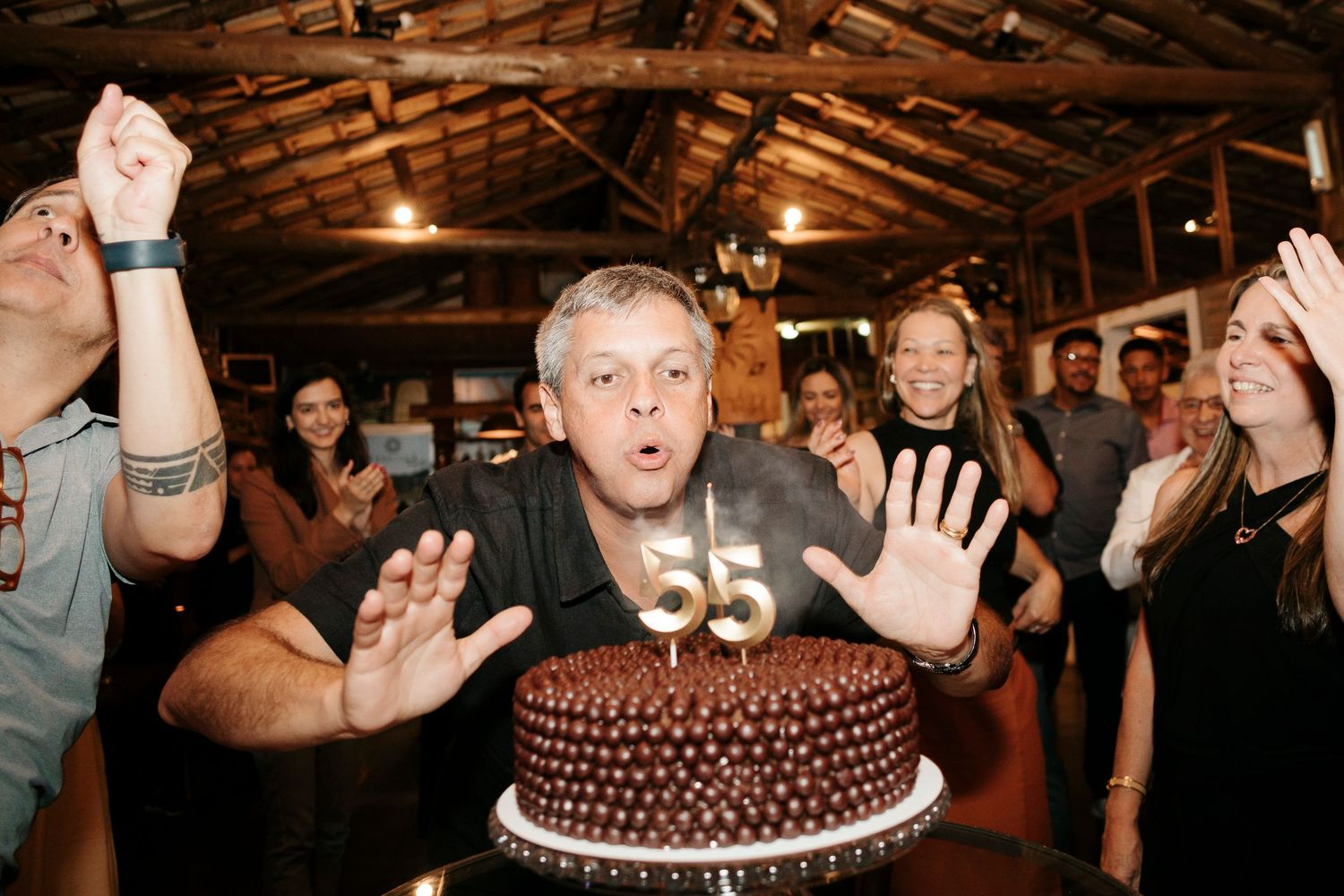 Person blowing out candles on chocolate birthday cake surrounded by party guests.