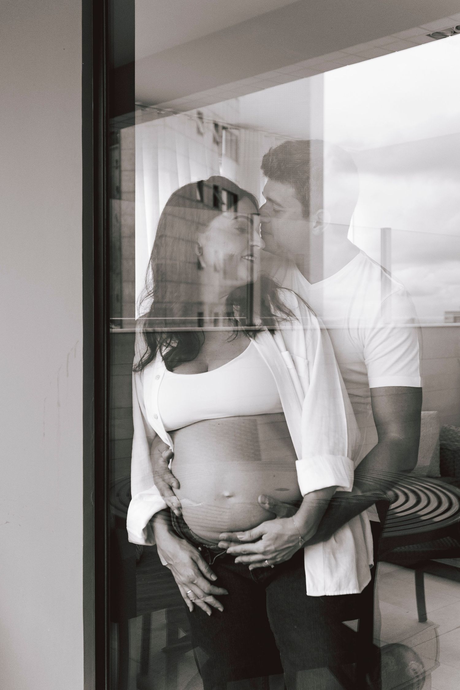 Artistic black and white photo of expectant couple embracing by window with reflected silhouette.