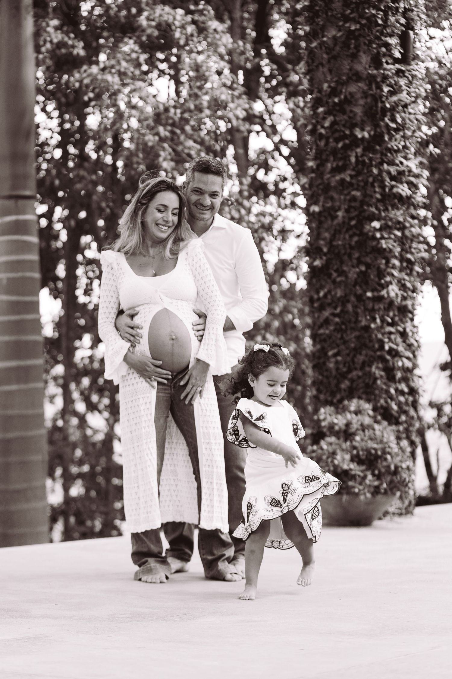 Black and white photo of a pregnant couple with their young daughter outdoors, surrounded by lush greenery.