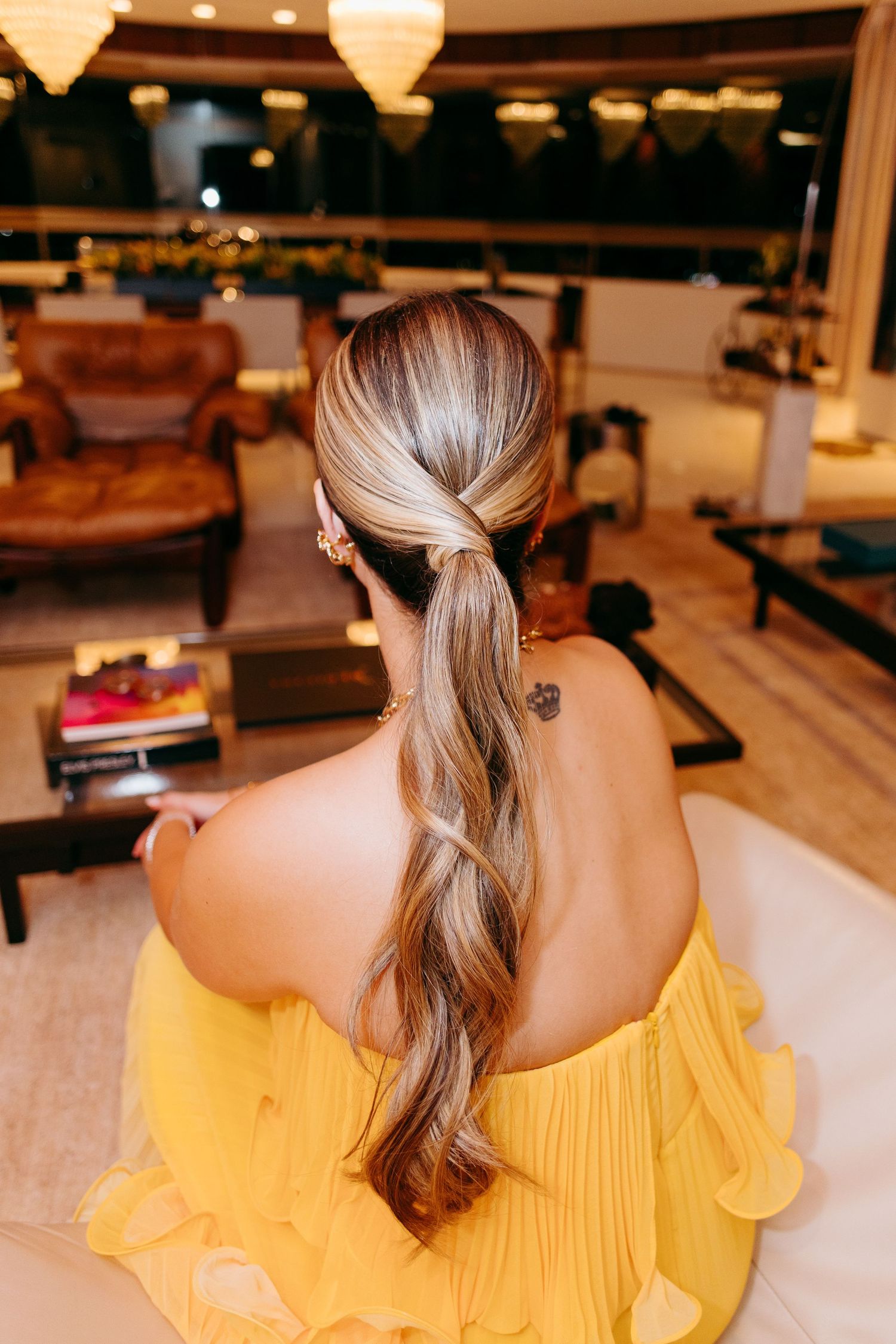 Back view of elegant hairstyle with long wavy ponytail and yellow off-shoulder dress in warm indoor setting.