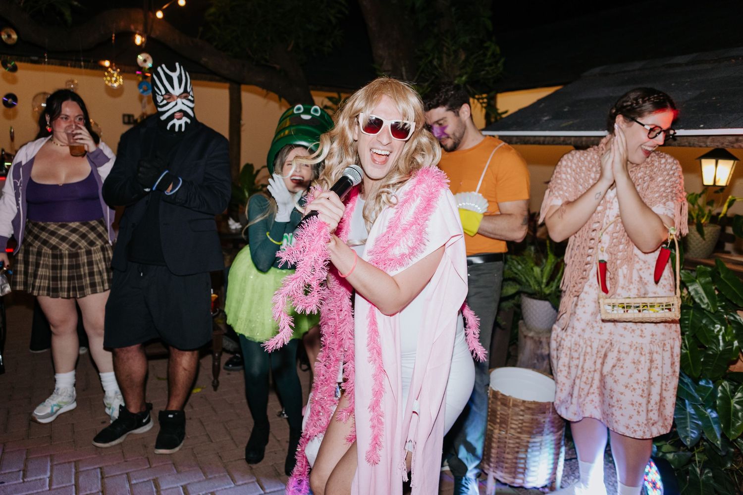 Party guests in colorful costumes dance and celebrate at an outdoor nighttime event with string lights.