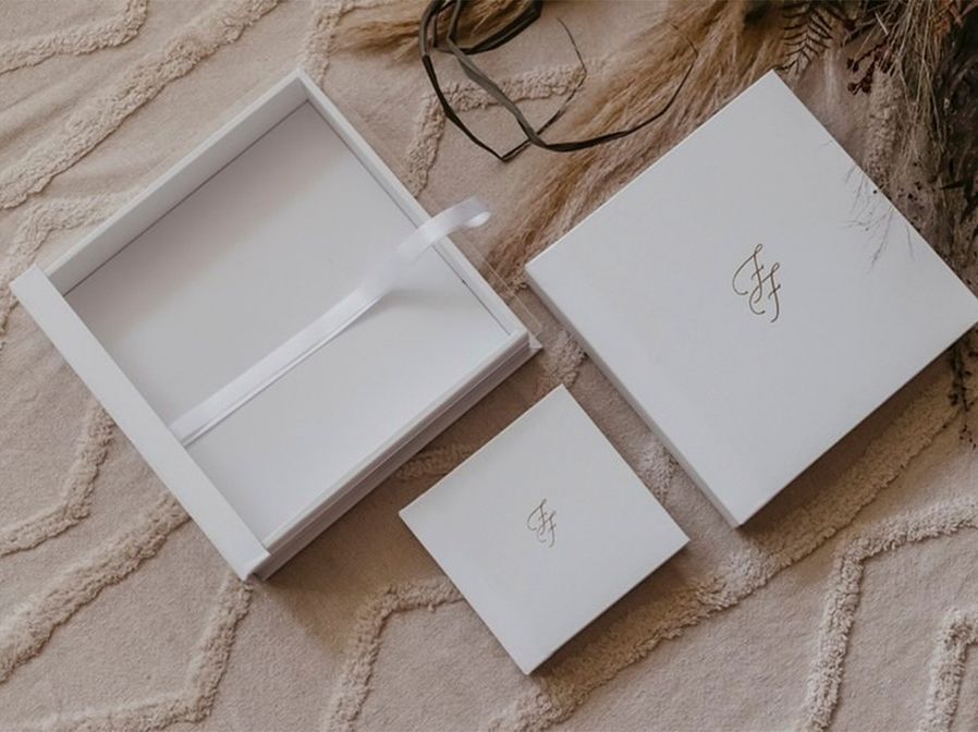 Elegant white gift boxes with ribbon and monogram logo displayed on a textured cream background.