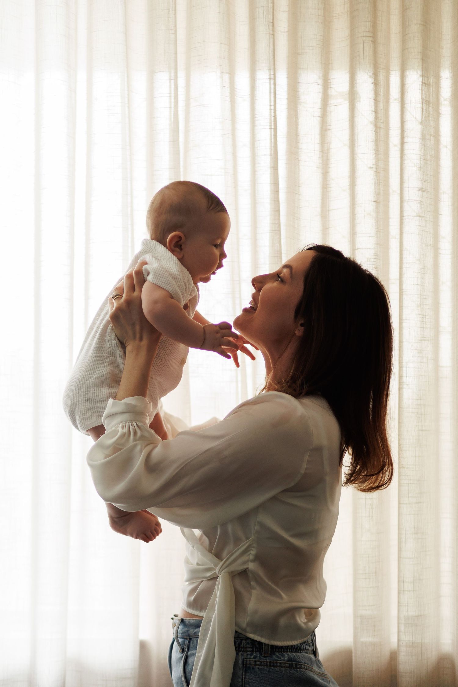 A mother lovingly holds her baby up near a bright window, both dressed in white, sharing a tender moment.