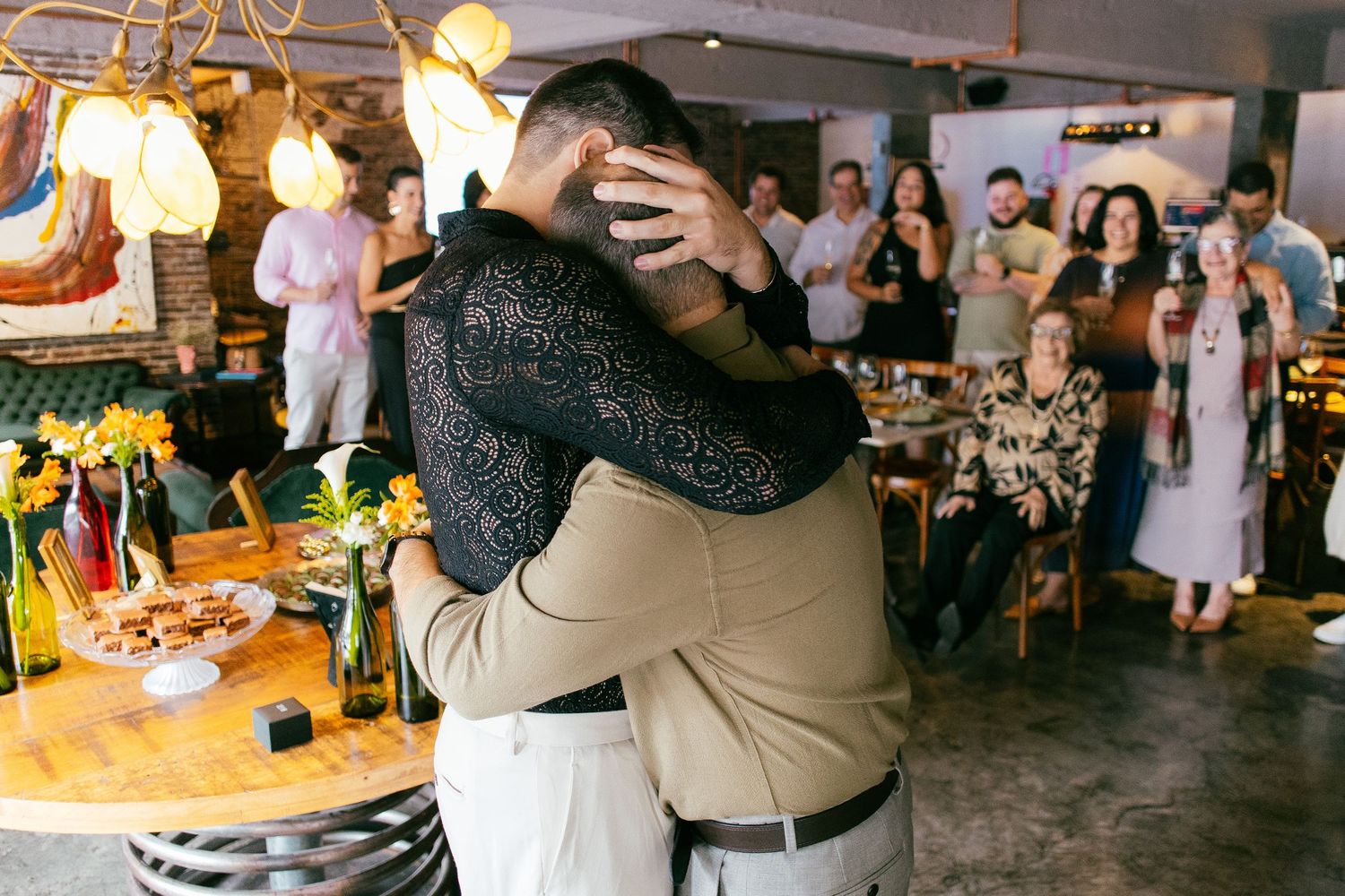 Two people share an emotional hug at a celebration event with guests watching in a warmly lit restaurant.