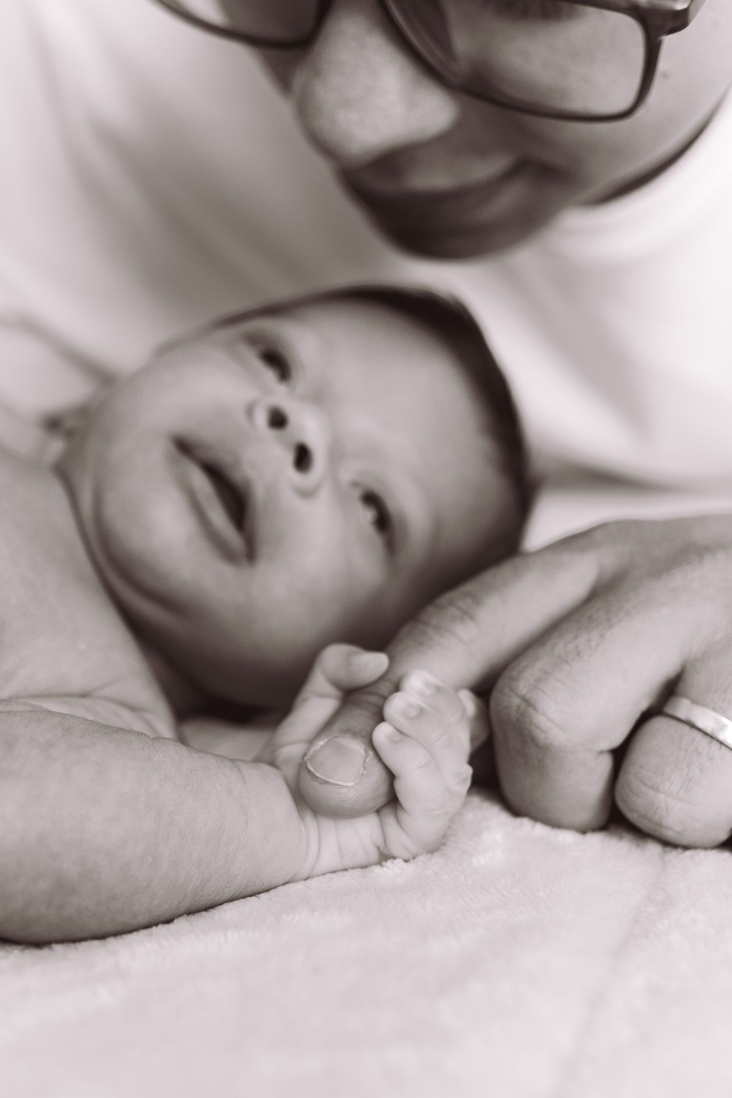Black and white photo of a newborn baby grasping an adult's finger while a parent smiles lovingly nearby.