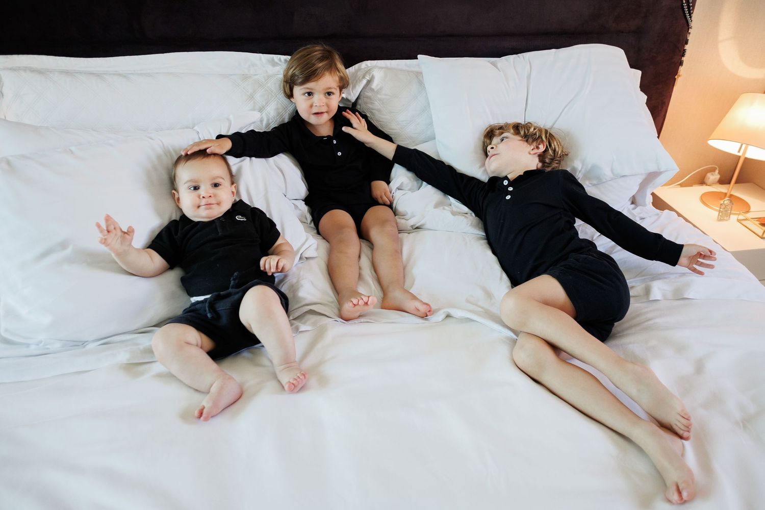 Three young siblings in black outfits lying together on a white bed in a cozy room.