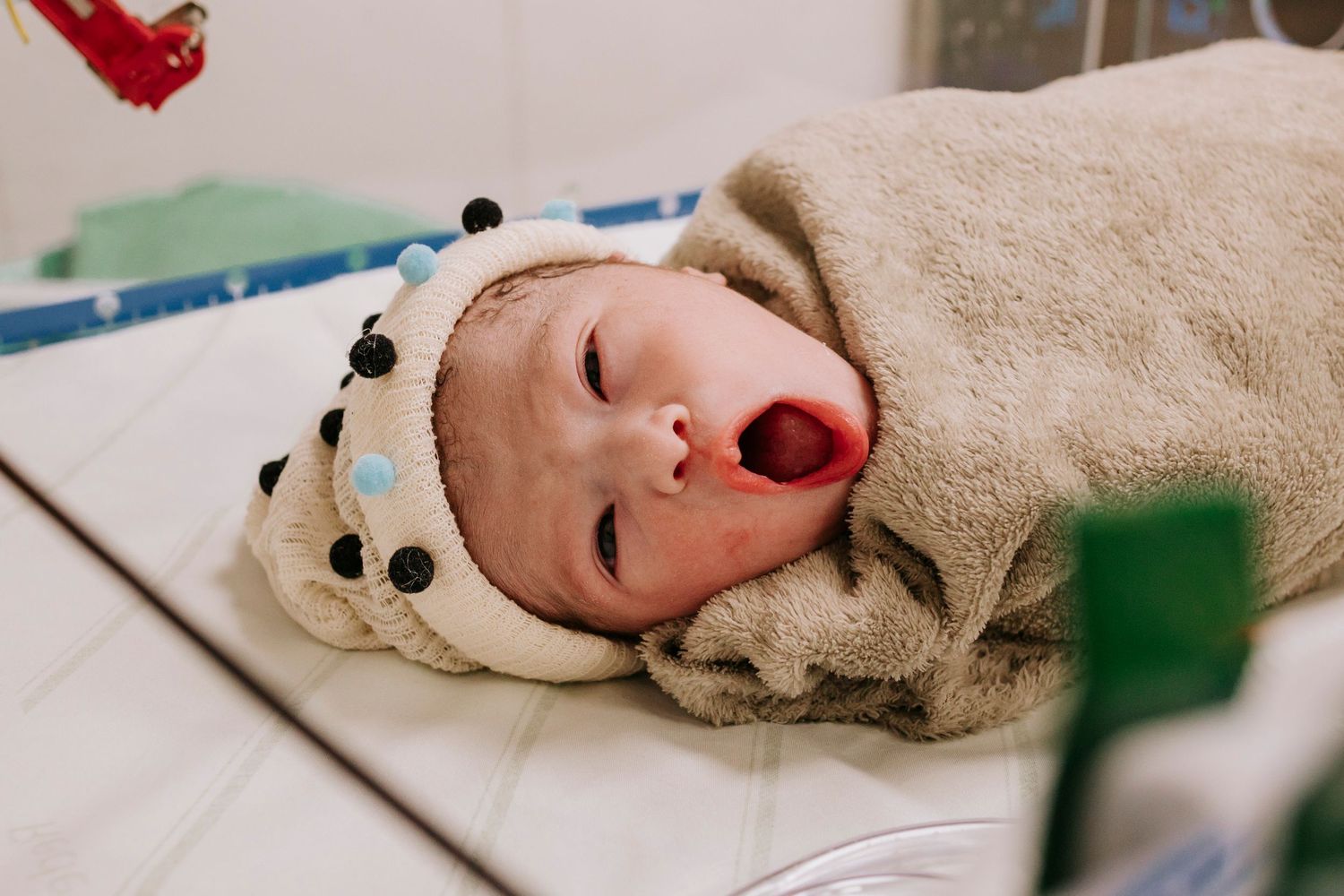 Newborn wrapped in beige blanket with polka dot hat yawning while lying on hospital bed.
