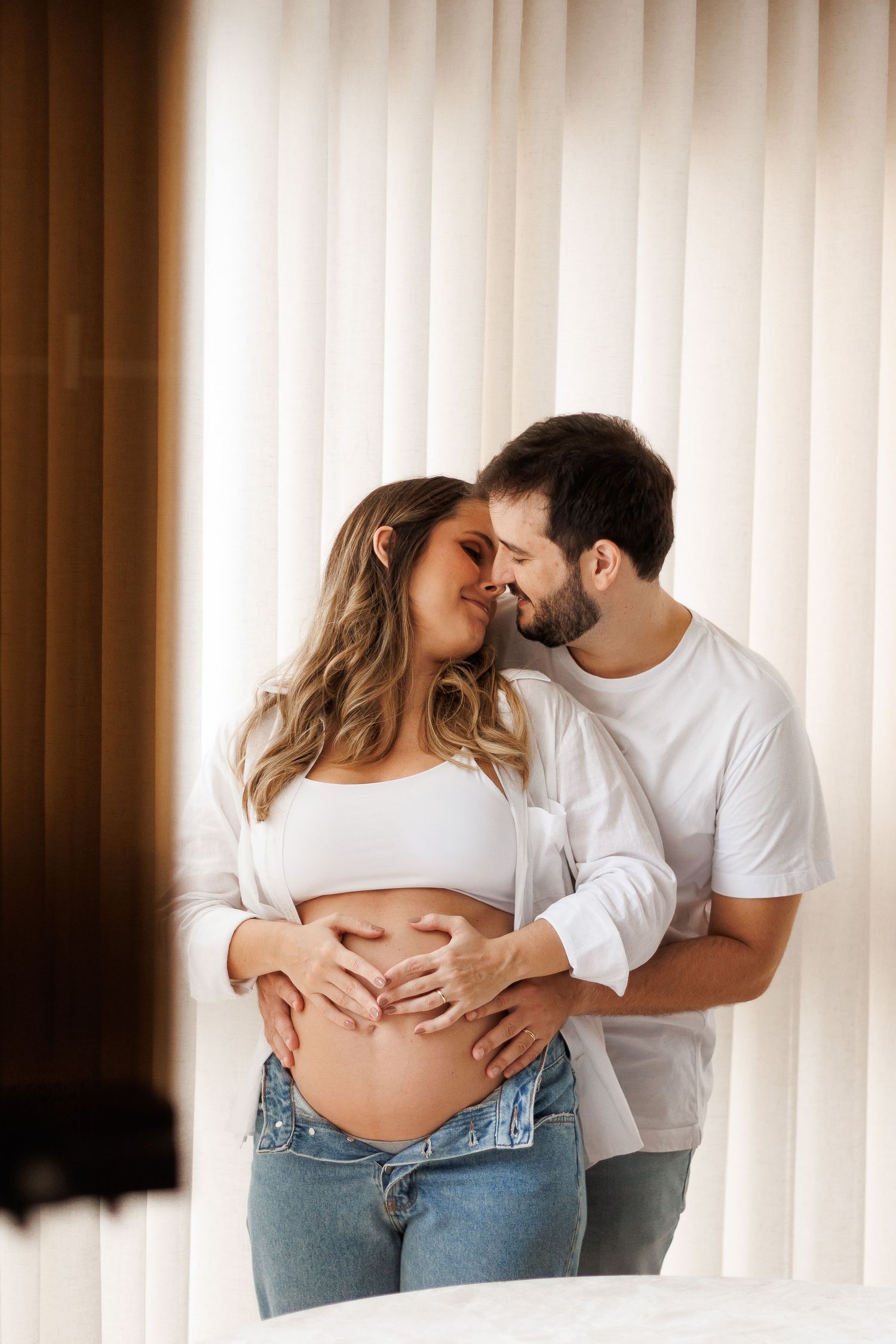Happy couple sharing a tender moment, man embracing pregnant woman making heart shape on baby bump.