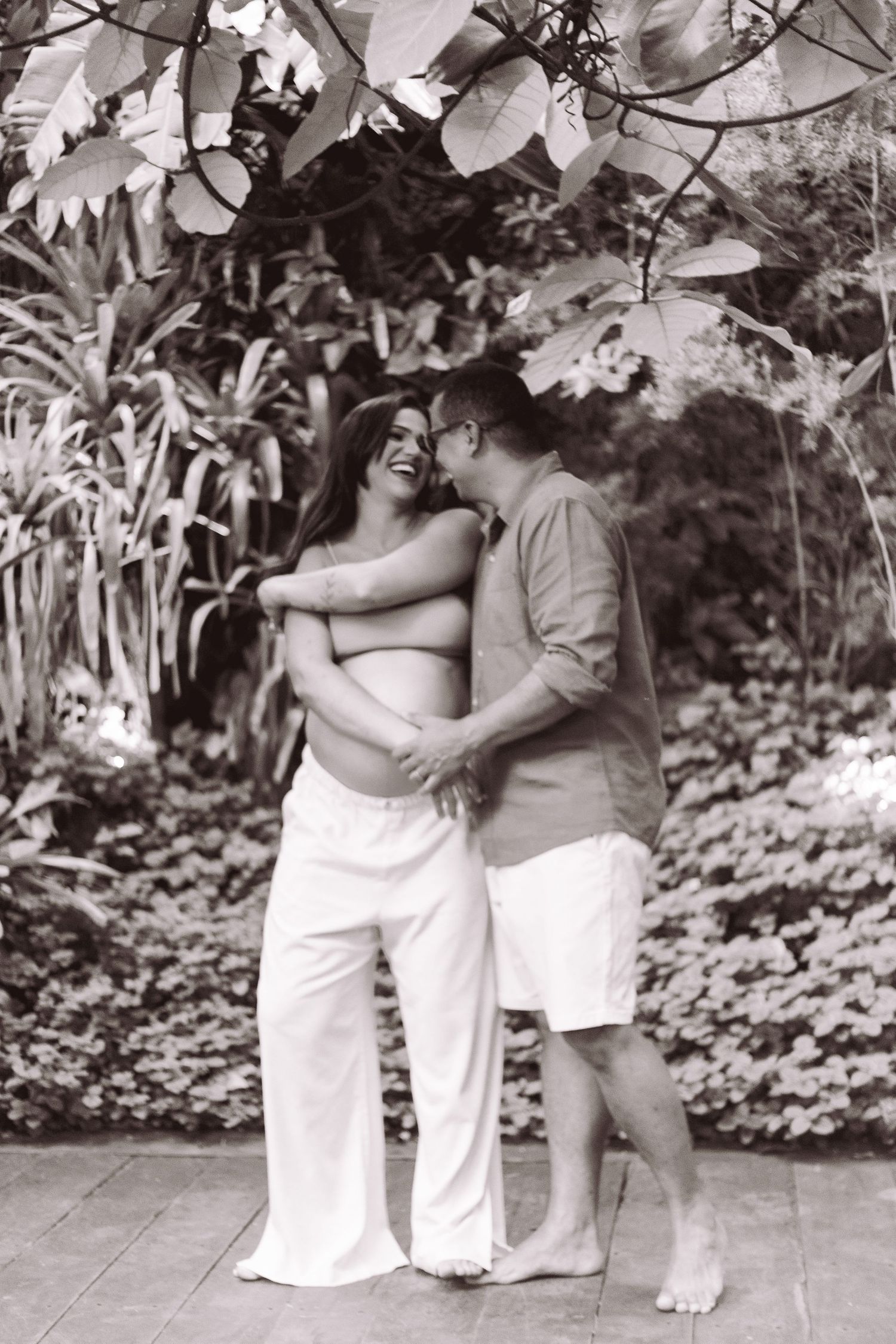 Black and white photo of happy couple embracing outdoors during maternity session surrounded by tropical plants.