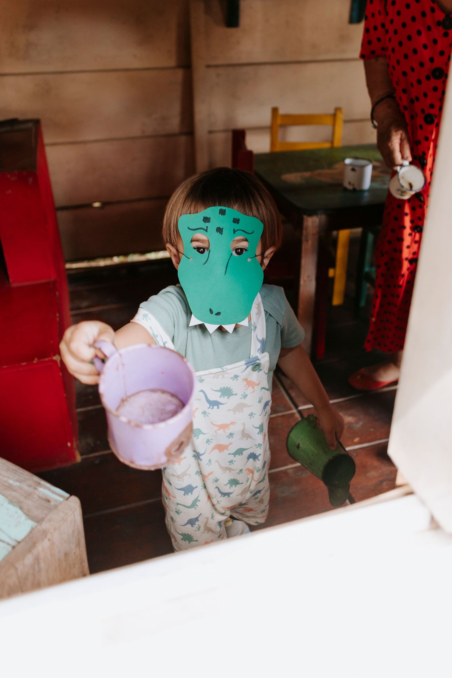 Child wearing dinosaur mask playing with toys in wooden playhouse.