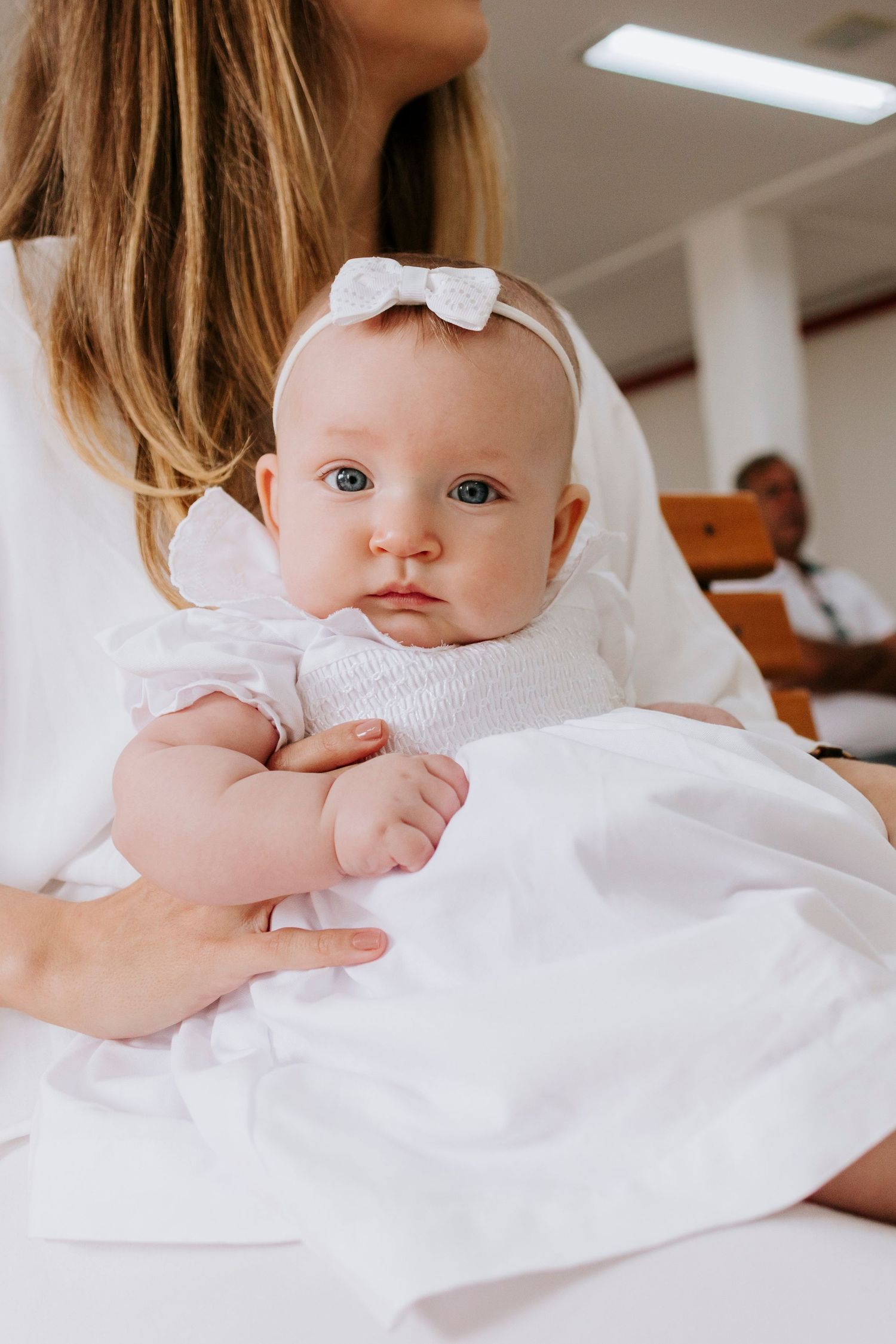 Baby in white dress and bow headband looking at camera.