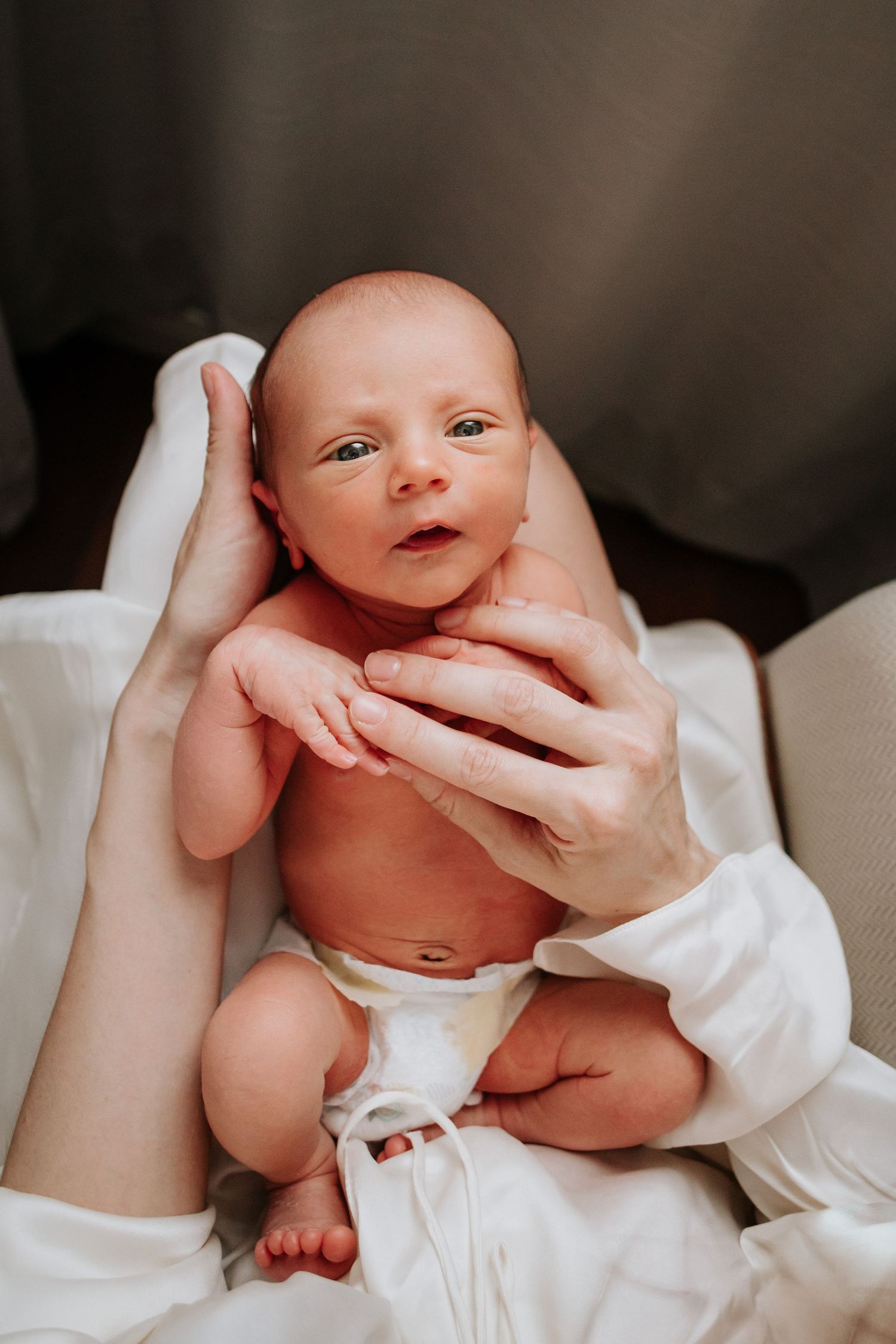 A small infant in a white diaper lays cradled in gentle hands against soft fabric.