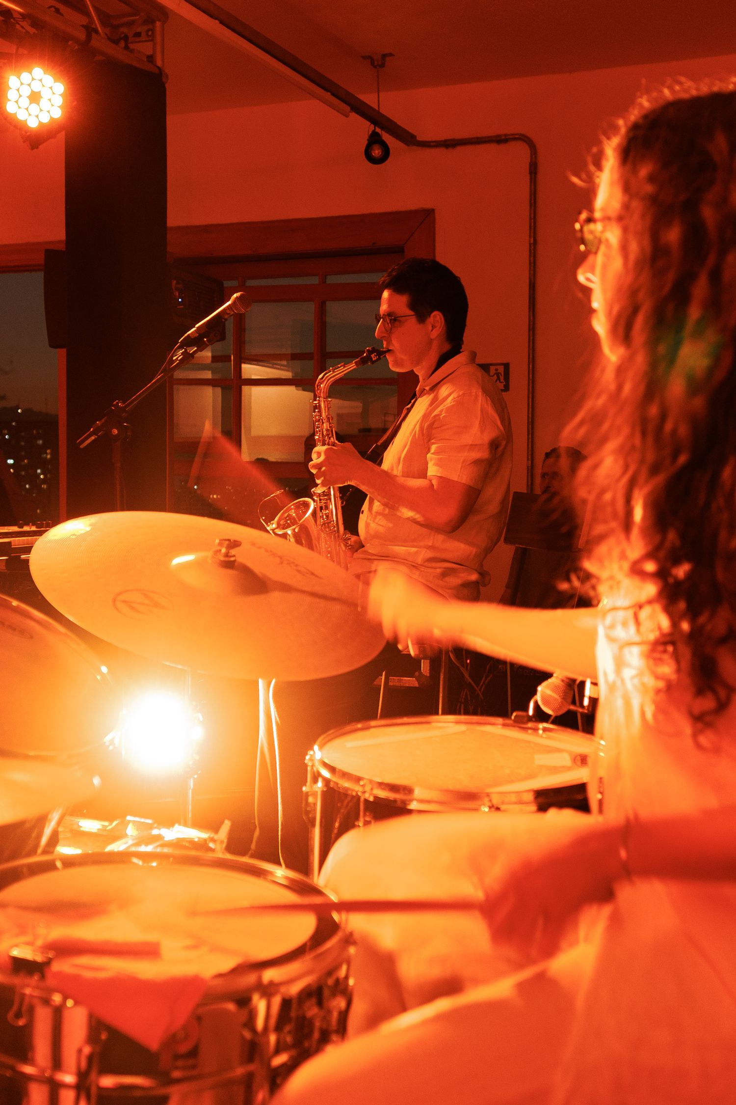 Musicians perform on stage in warm orange lighting with a saxophone player and drummer visible during a live jazz performance.