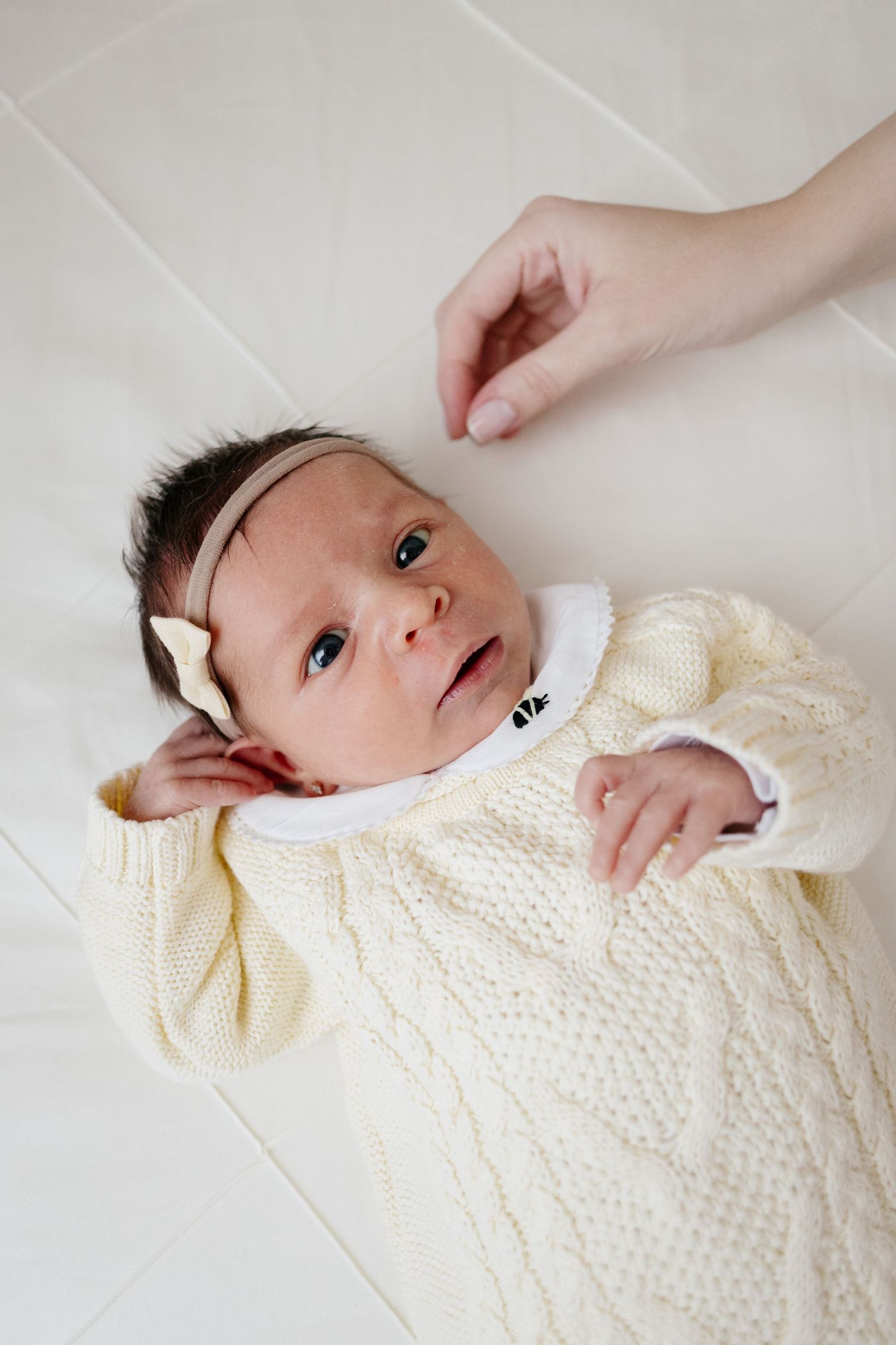 Newborn baby girl in cream knit sweater and bow headband lying on white surface with mother's hand nearby.