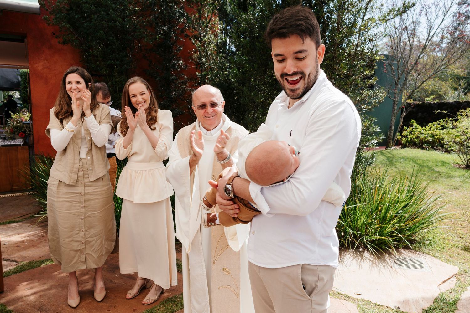 A group wearing white and beige outfits poses together in a garden setting on a sunny day.