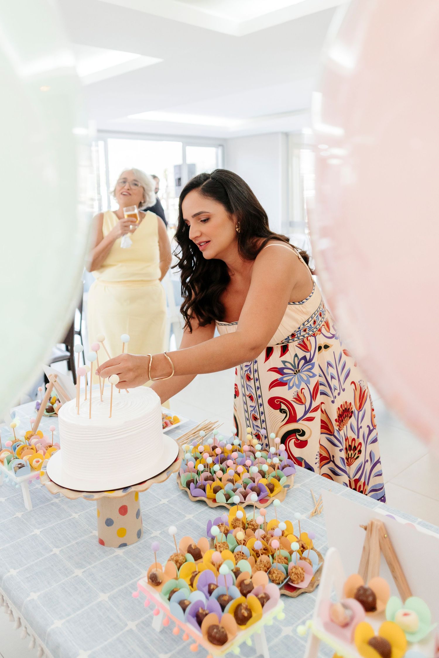 A person in a floral dress arranges colorful mini donuts on a table next to a white cake display.