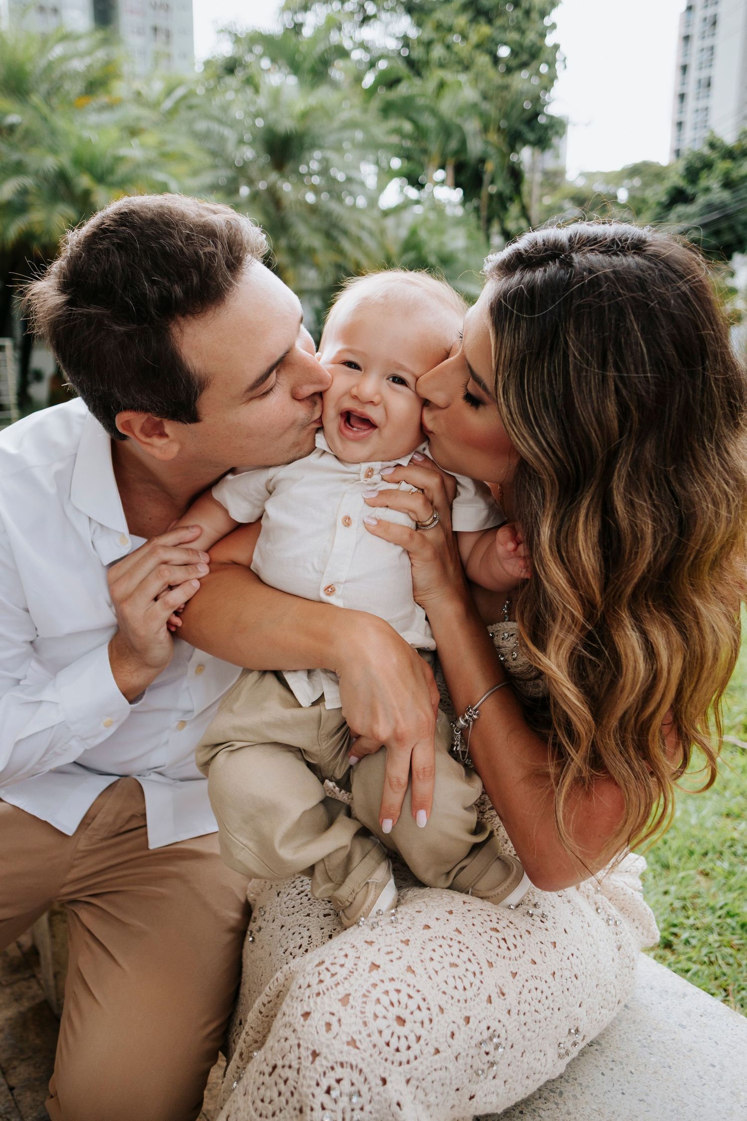 Intimate family moment outdoors with a laughing baby being kissed by parents on a sunny day.