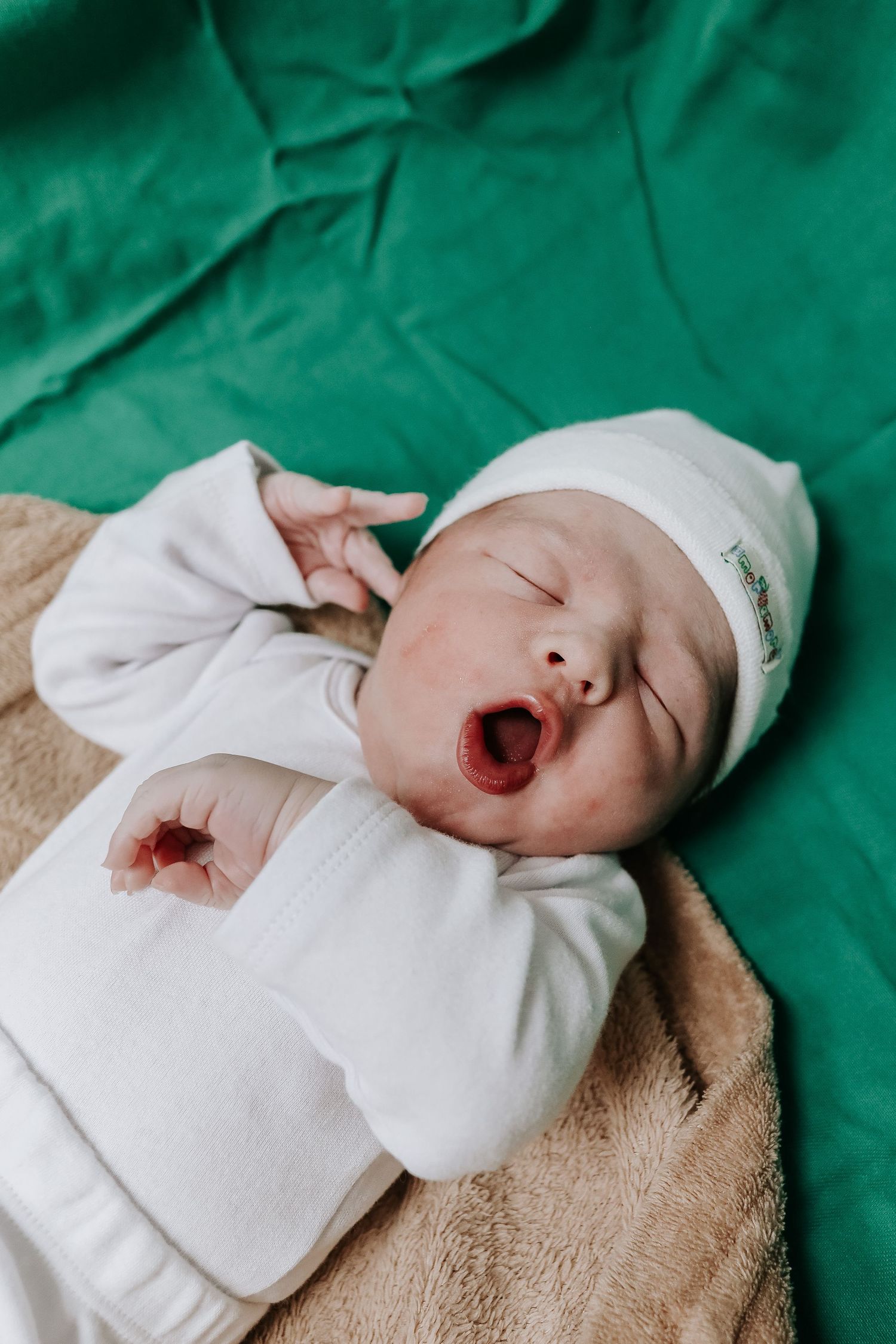 A newborn baby in white clothing yawns while lying on a green hospital blanket.