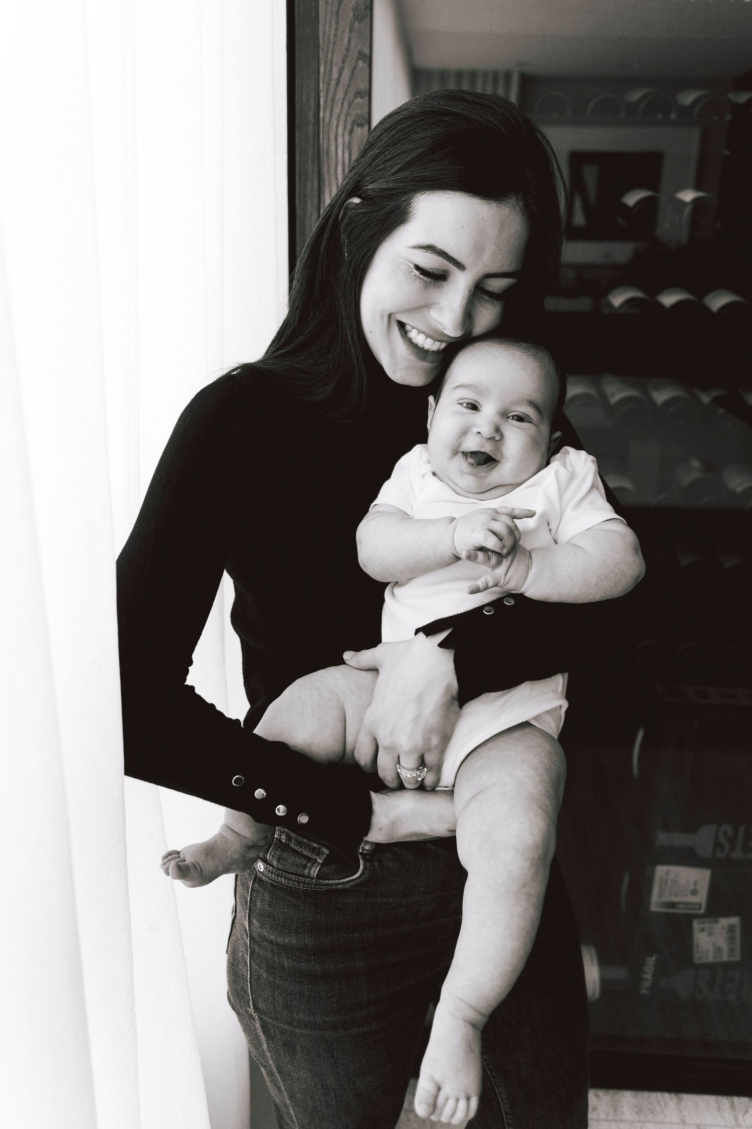Mother in black sweater holding smiling baby in white onesie by window.
