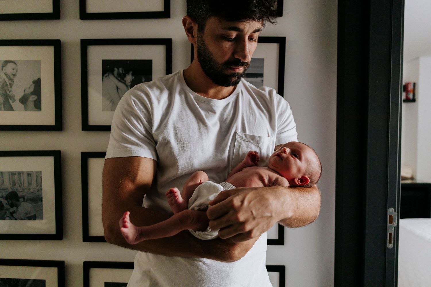 Parent in white t-shirt holding sleeping newborn against wall with framed photos.