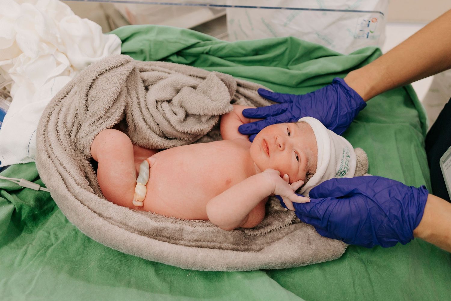 Healthcare workers in blue gloves examining newborn baby wrapped in towel.