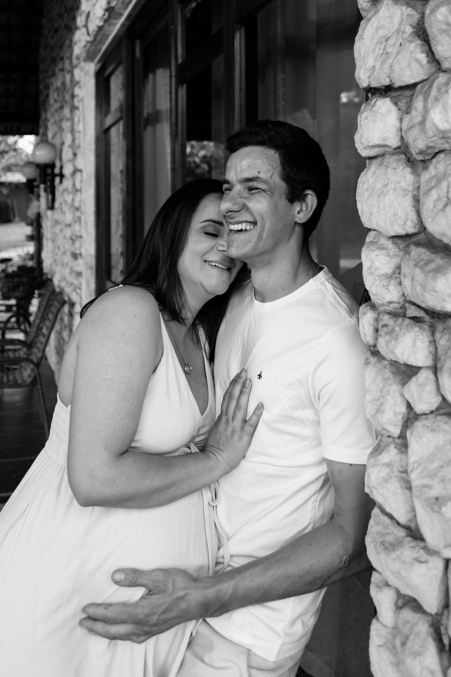 Black and white portrait of expecting couple laughing and embracing by stone wall.