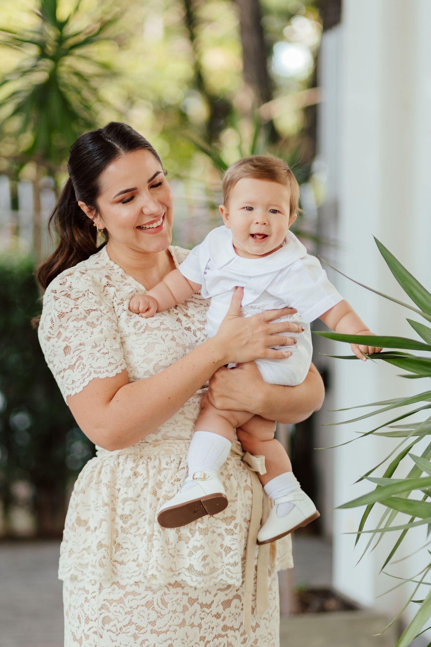 A woman in a white lace dress shares joyful moments with a baby outdoors near tropical plants.