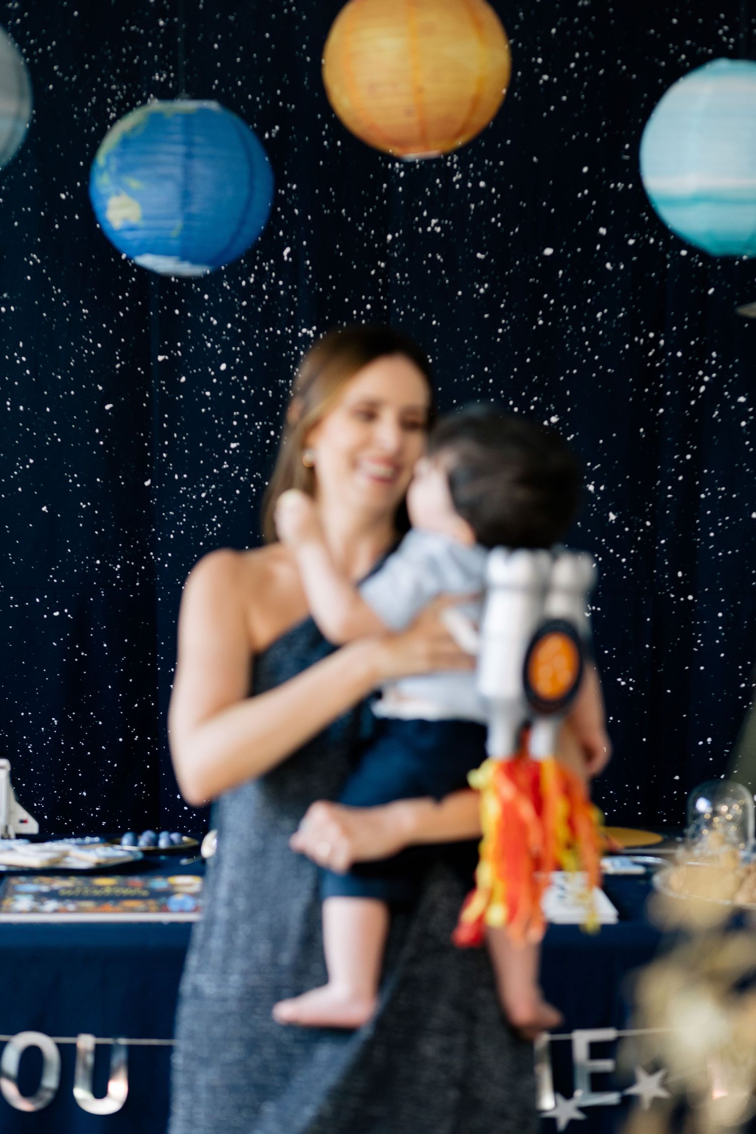 Woman holding baby at space-themed birthday party with planet lanterns and starry backdrop.