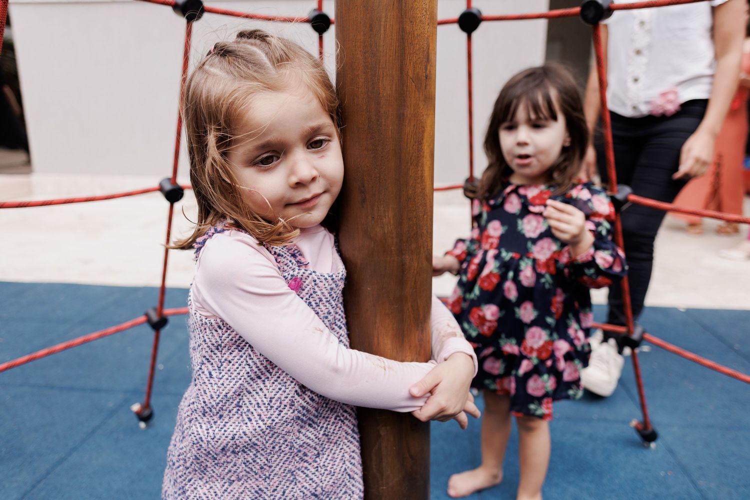 Two young children play at an indoor climbing structure with ropes and poles on a blue padded floor.
