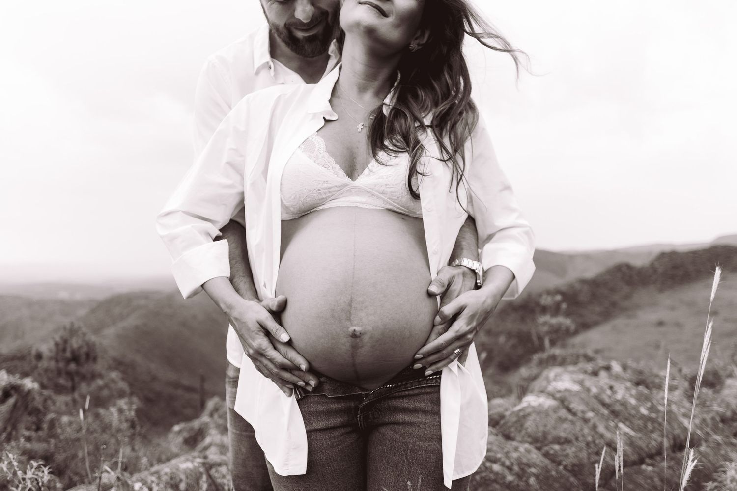 Black and white photo of a pregnant couple outdoors, man embracing woman from behind, both cradling her baby bump.