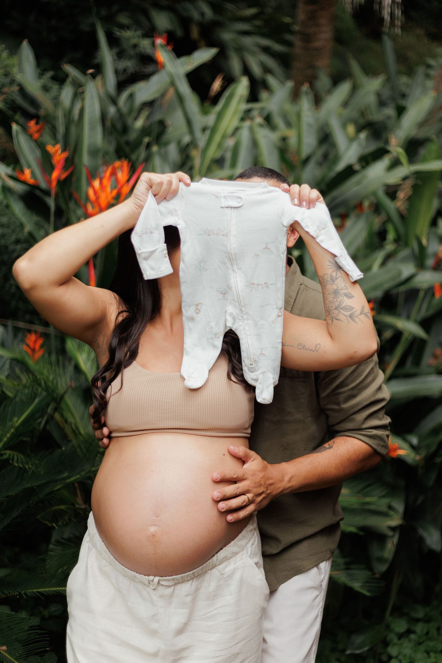 Pregnant couple holding up tiny white baby onesie in front of faces surrounded by tropical garden foliage.
