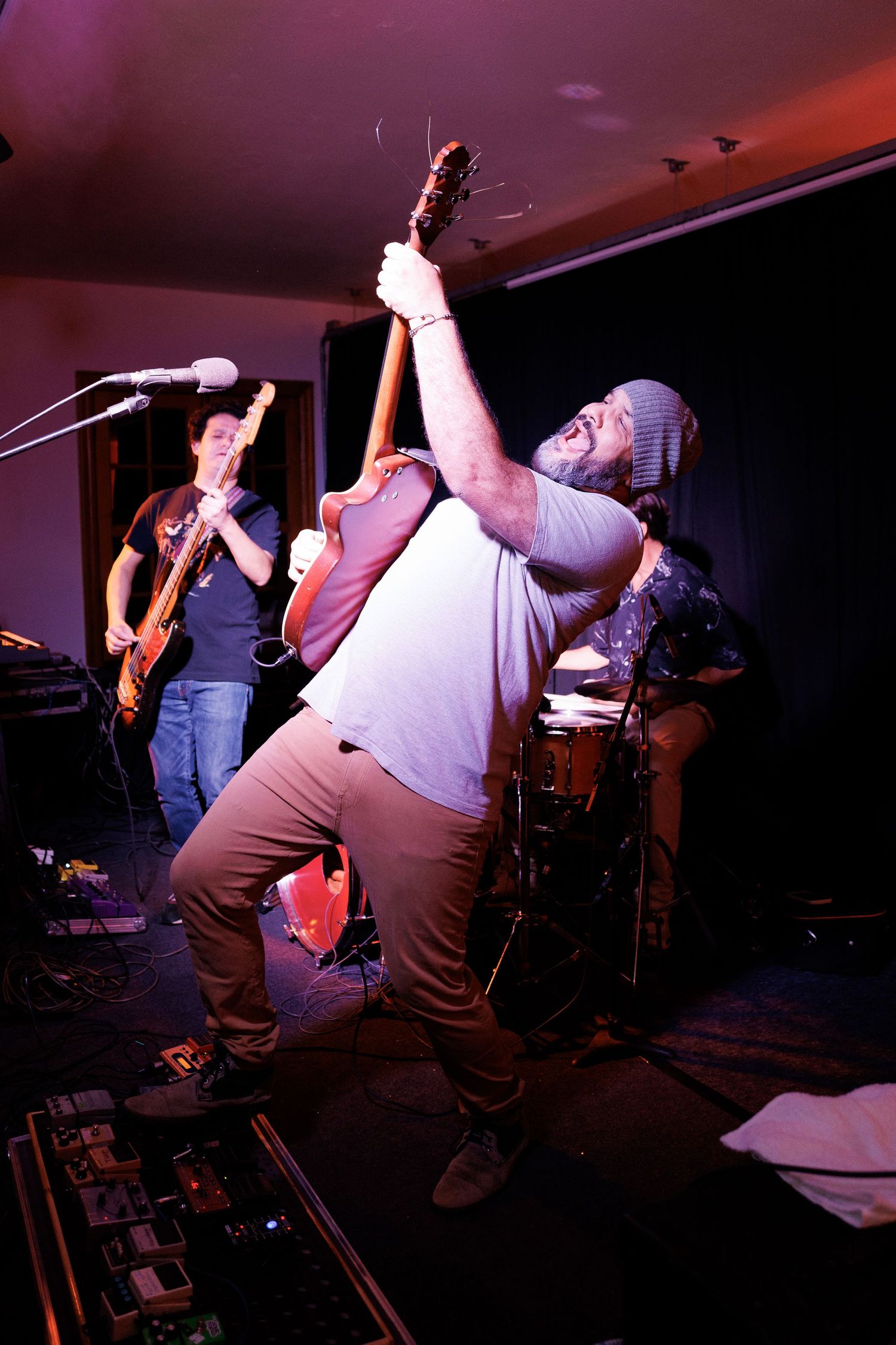 Energetic performer holds microphone while singing passionately on stage with band members visible in background.
