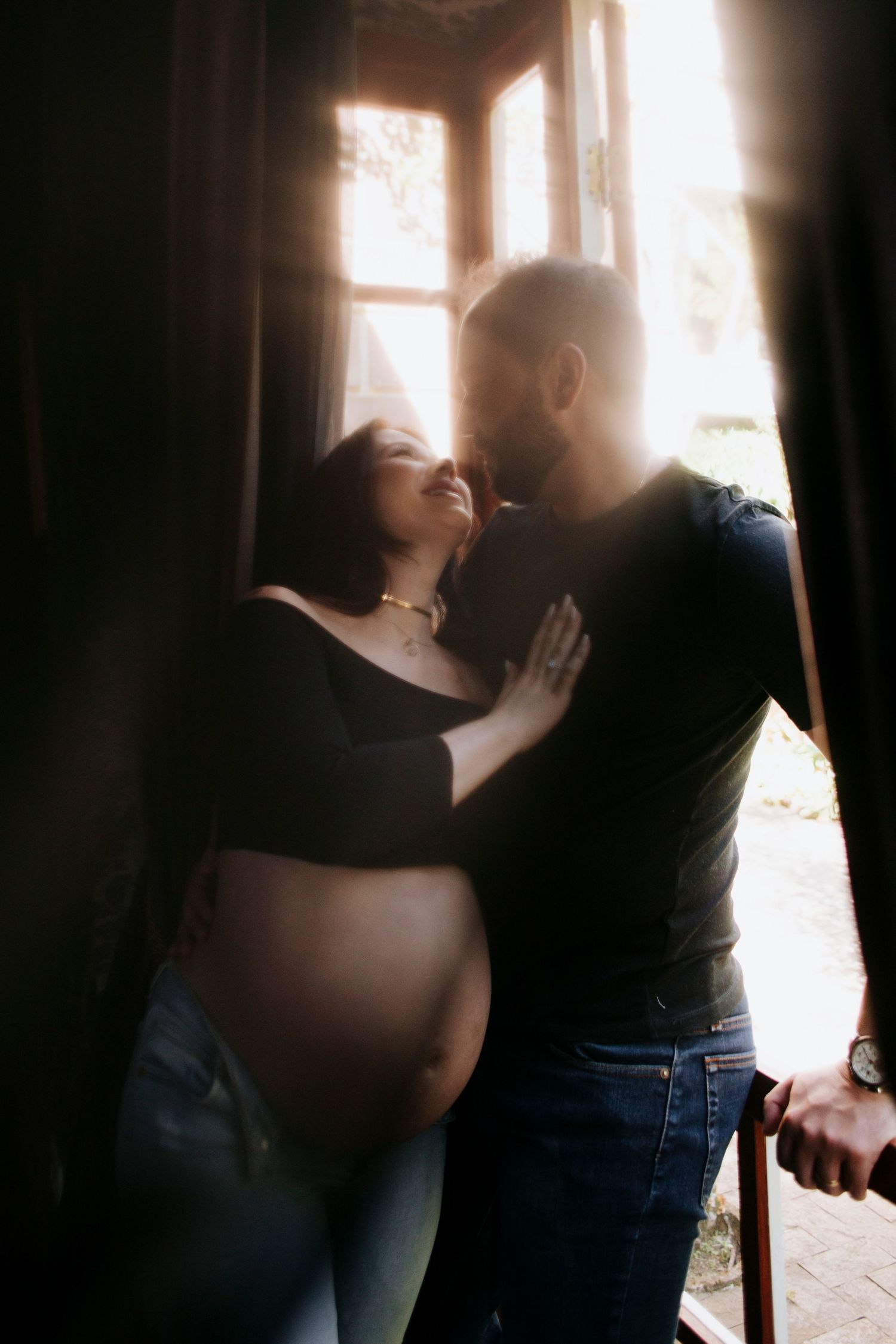 Romantic maternity photo of couple embracing near window with soft lighting.