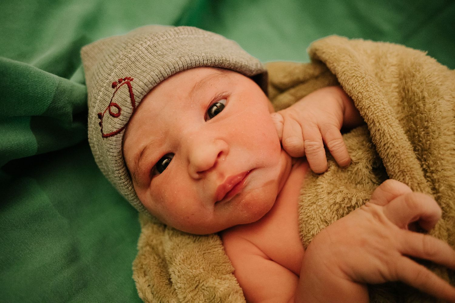 Newborn baby wearing a knit hat smiles while wrapped in a brown blanket against green background.