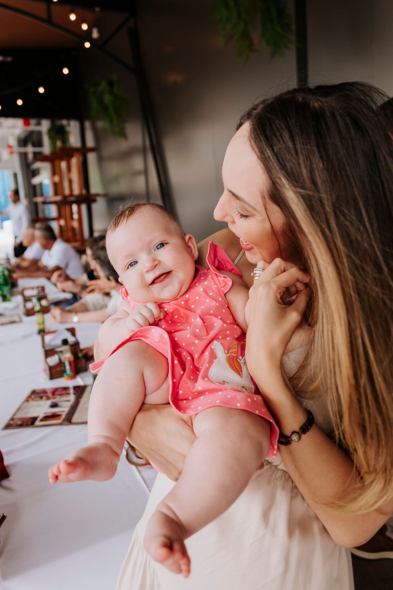 Mother holding smiling baby in coral romper at restaurant gathering.
