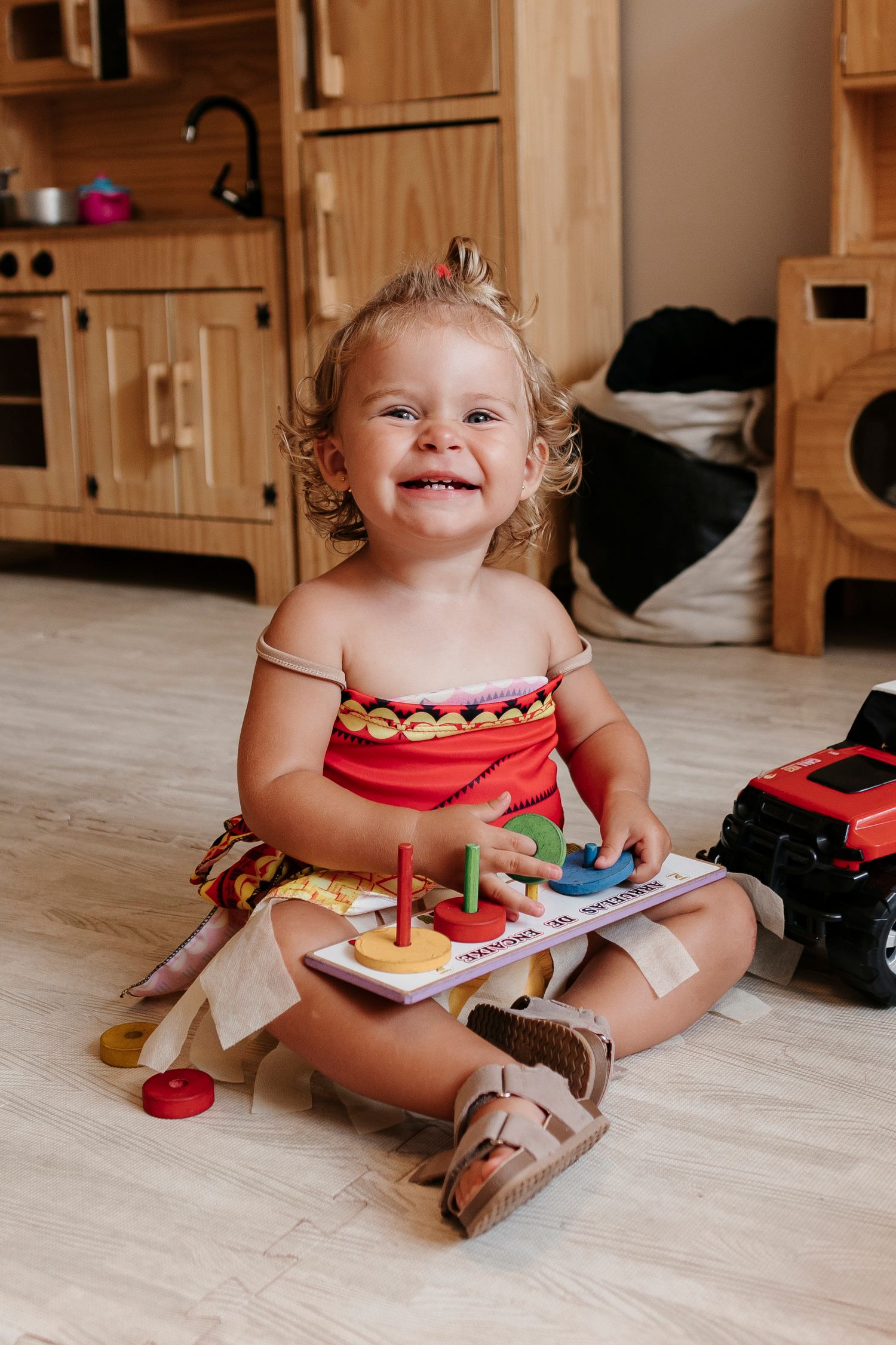Smiling toddler playing with colorful toys while sitting on wooden floor in play kitchen area.