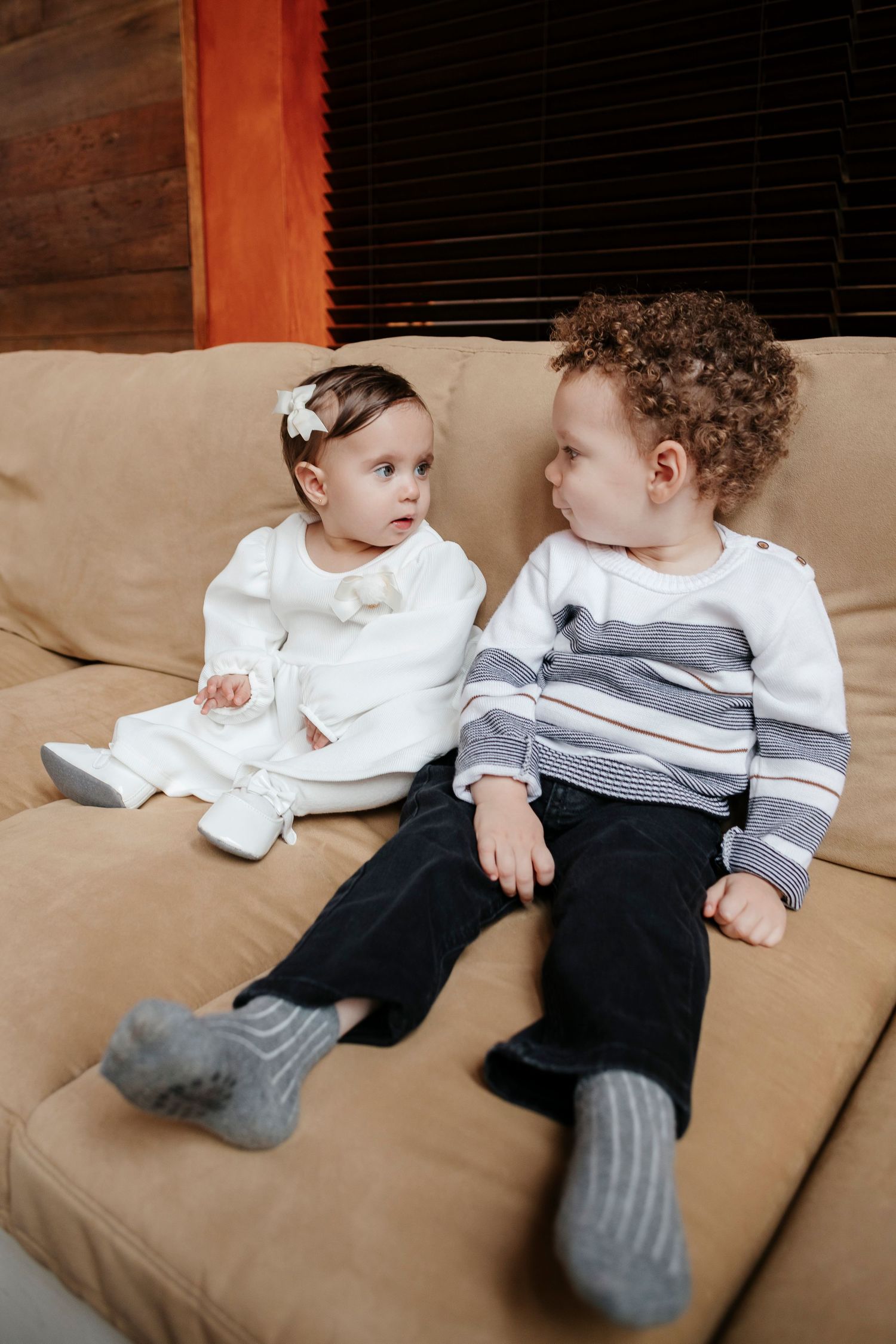 Two young children in formal wear sitting on beige couch looking at each other.