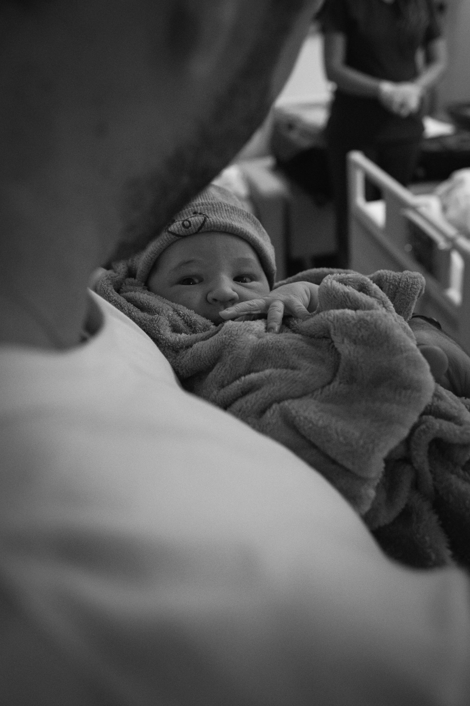 Black and white image of a swaddled newborn wearing a knit cap in a hospital setting.