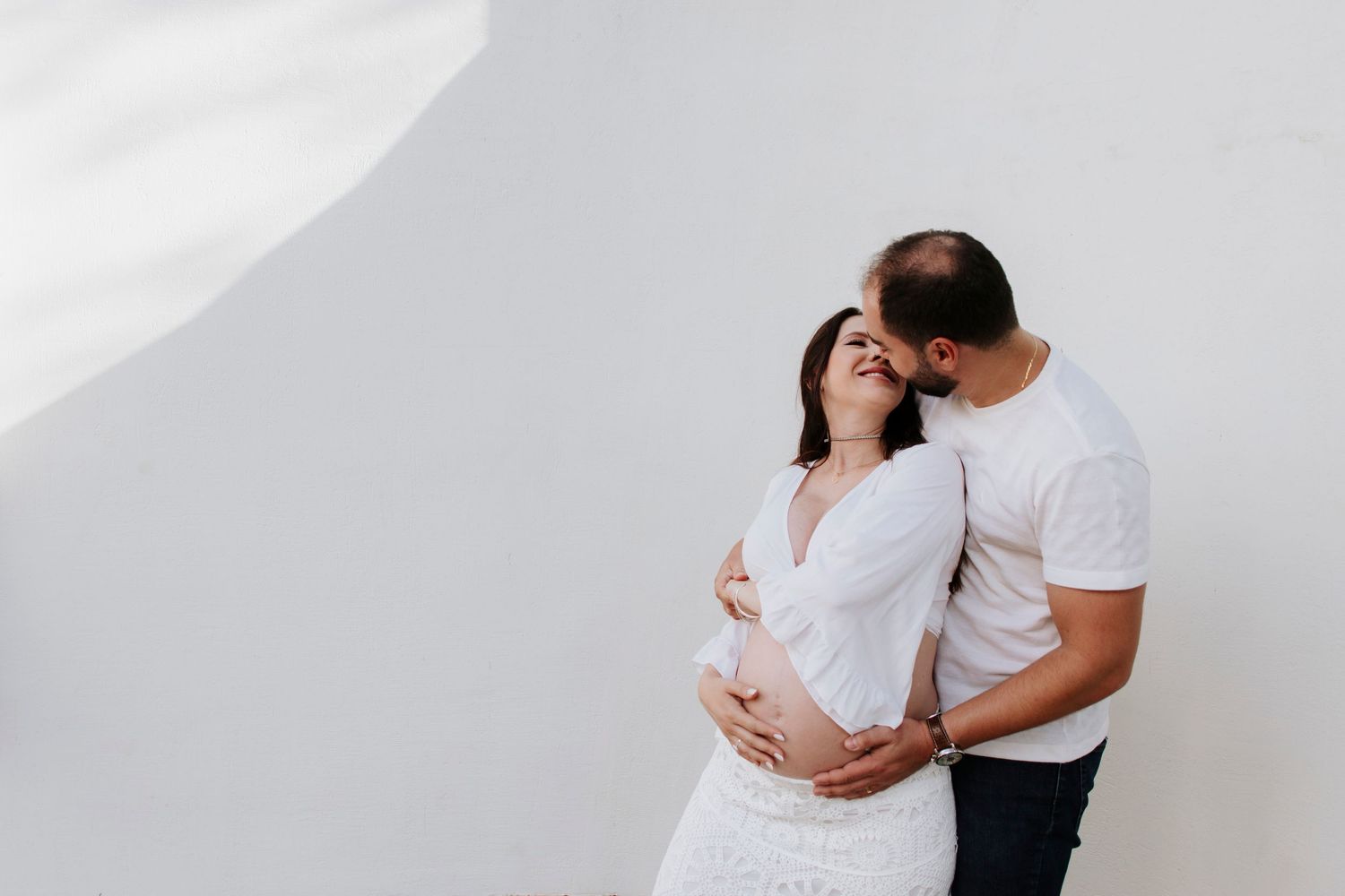 Expecting couple in white embracing against neutral wall in minimalist maternity photo.