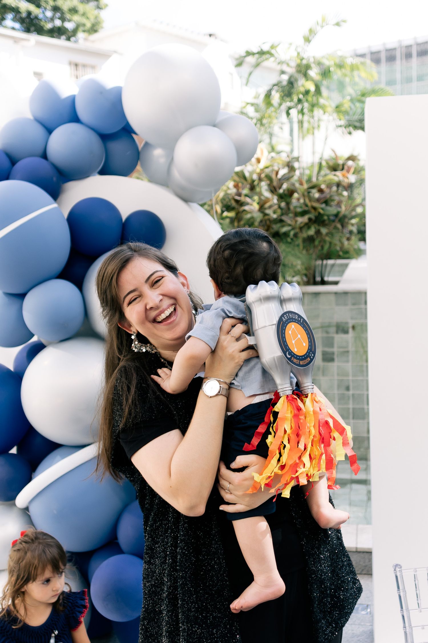 A joyful woman holds a baby in a rocket costume at a birthday party with blue and white balloon decorations.