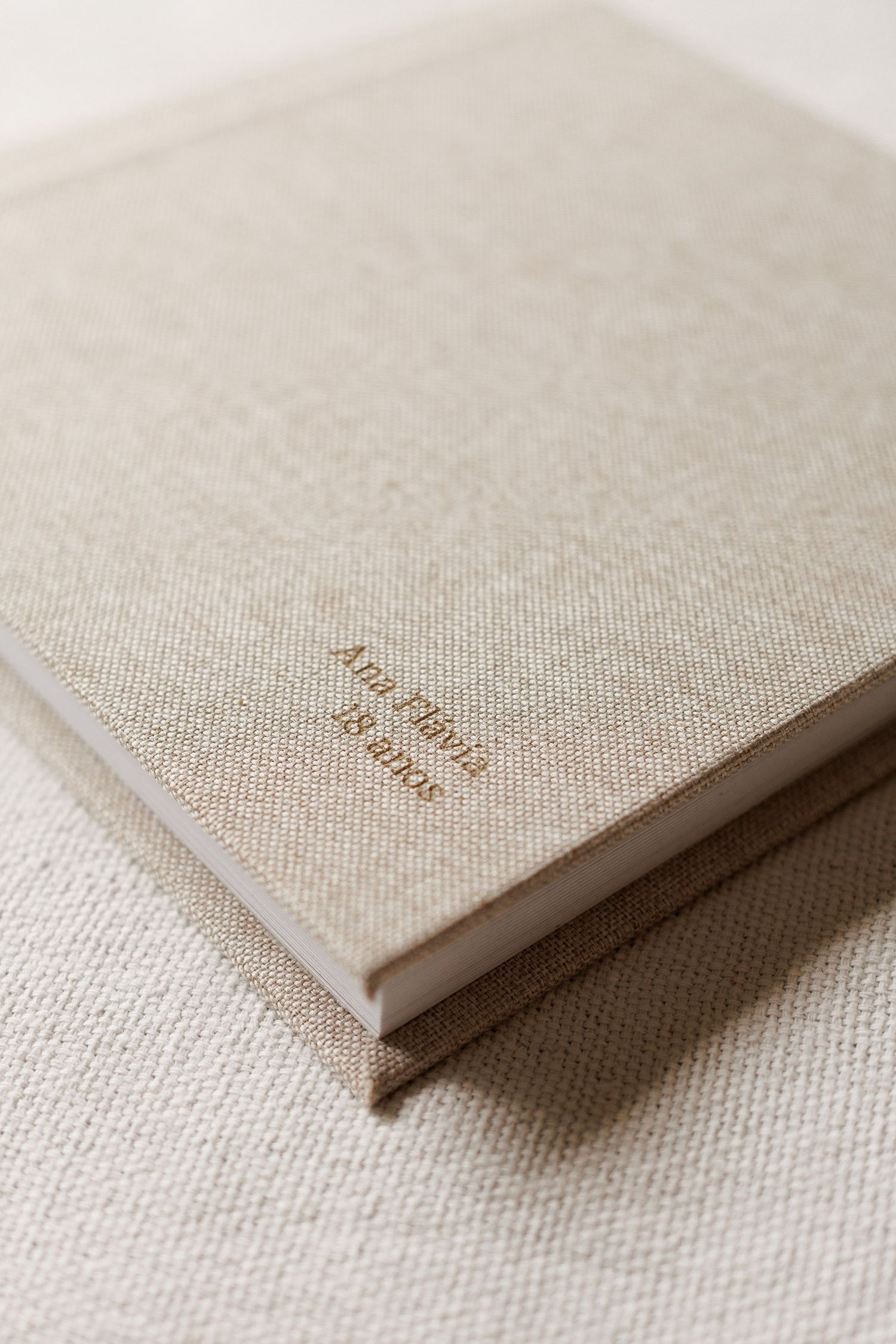 Elegant linen-covered journal with gold foil text reading Ana Flavia 18 anos, resting on a neutral fabric surface.