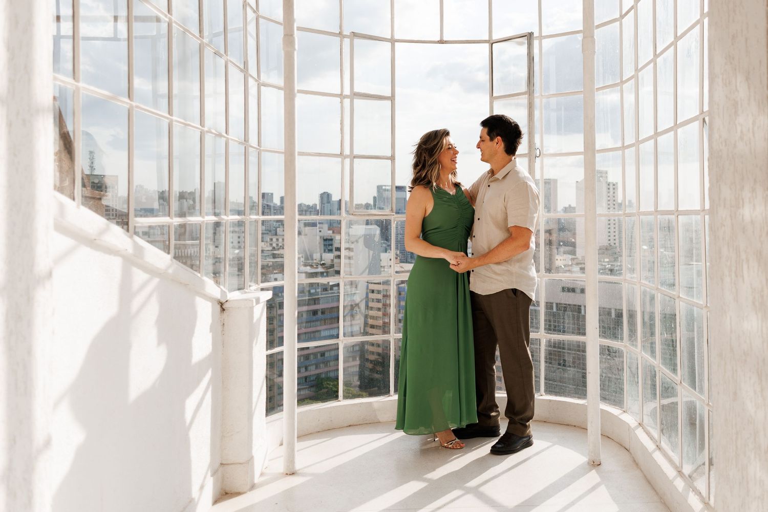 A couple embraces in an elegant curved glass observatory overlooking a cityscape on a sunny afternoon.