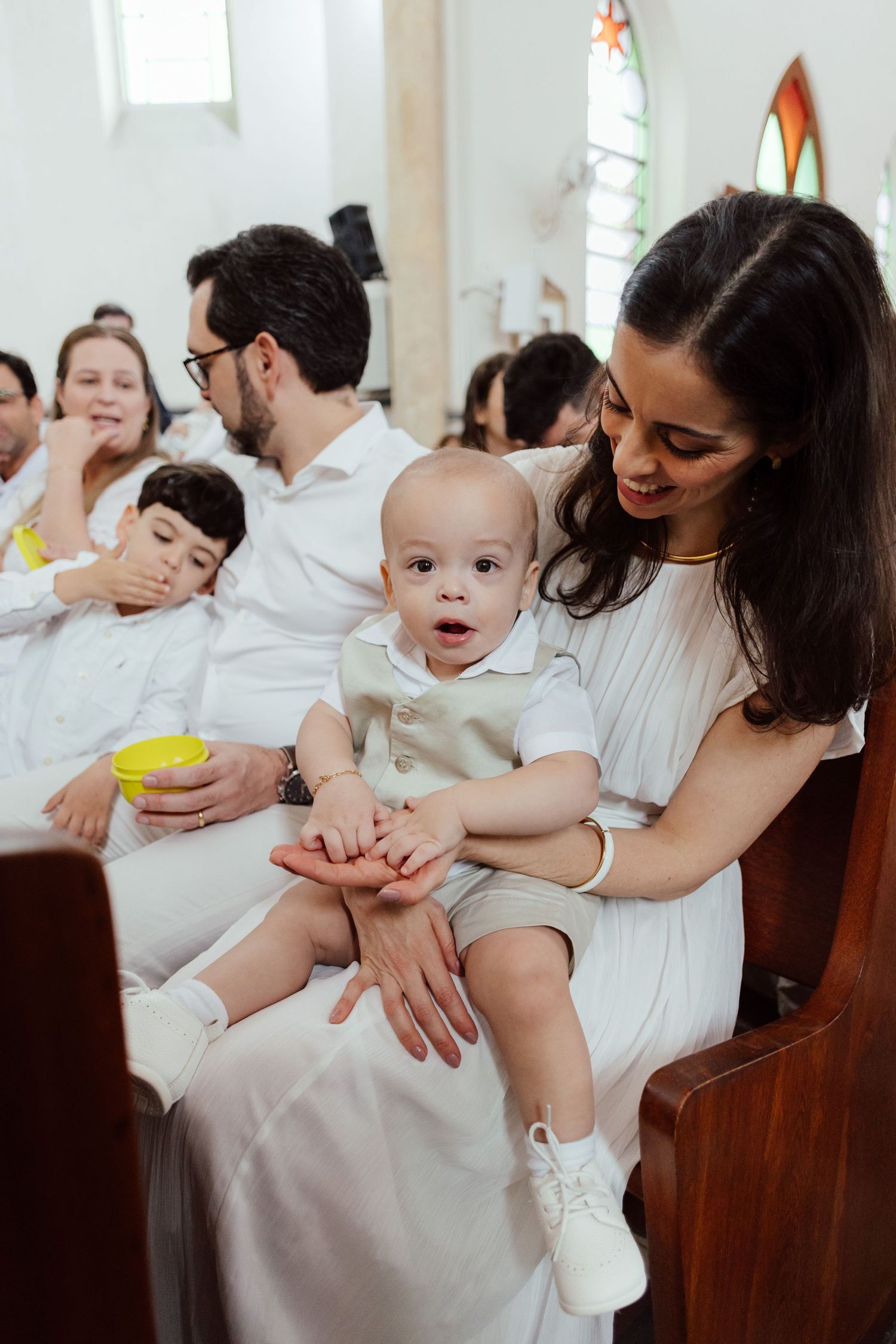 Multiple people dressed in white sit in church pews while interacting with young children during a religious ceremony.