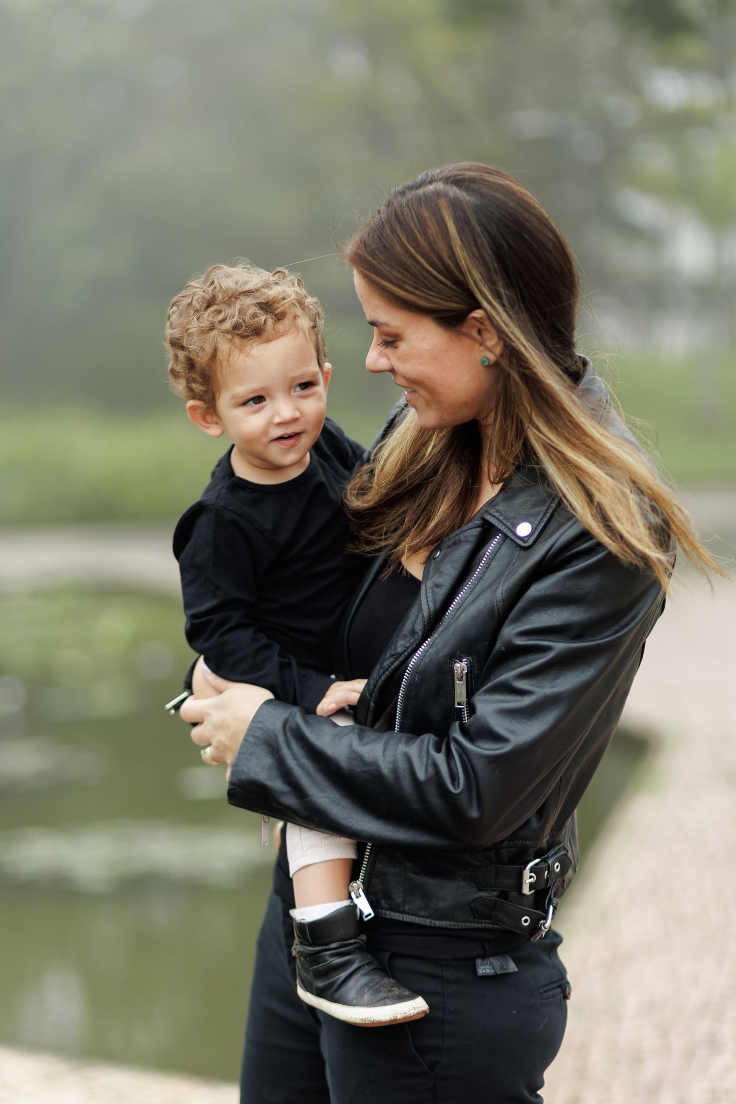 A mother in a black leather jacket lovingly holds her curly-haired toddler near a misty pond.