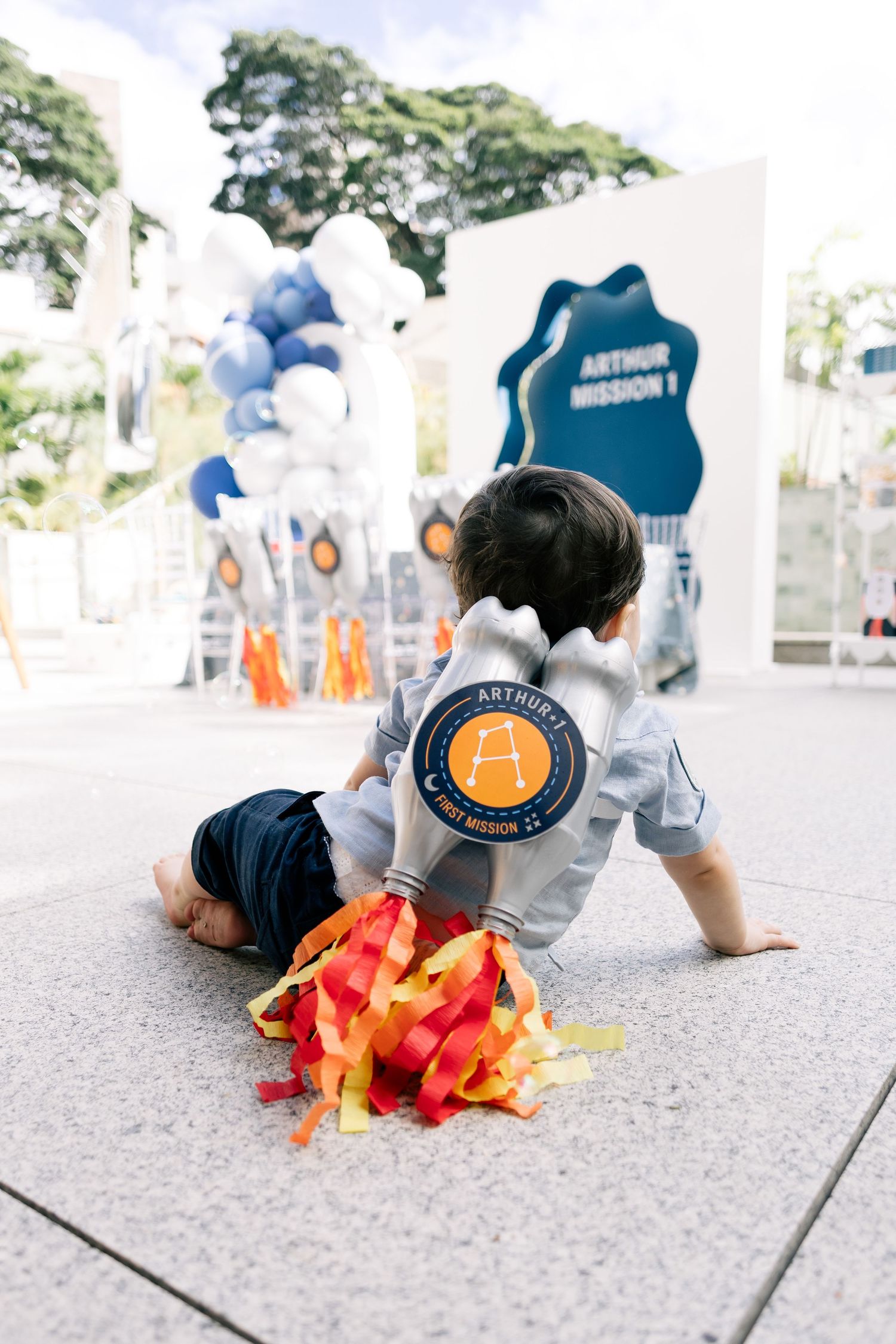 Baby boy in personalized astronaut jacket crawls toward colorful ribbons at Arthur's first birthday space-themed party.