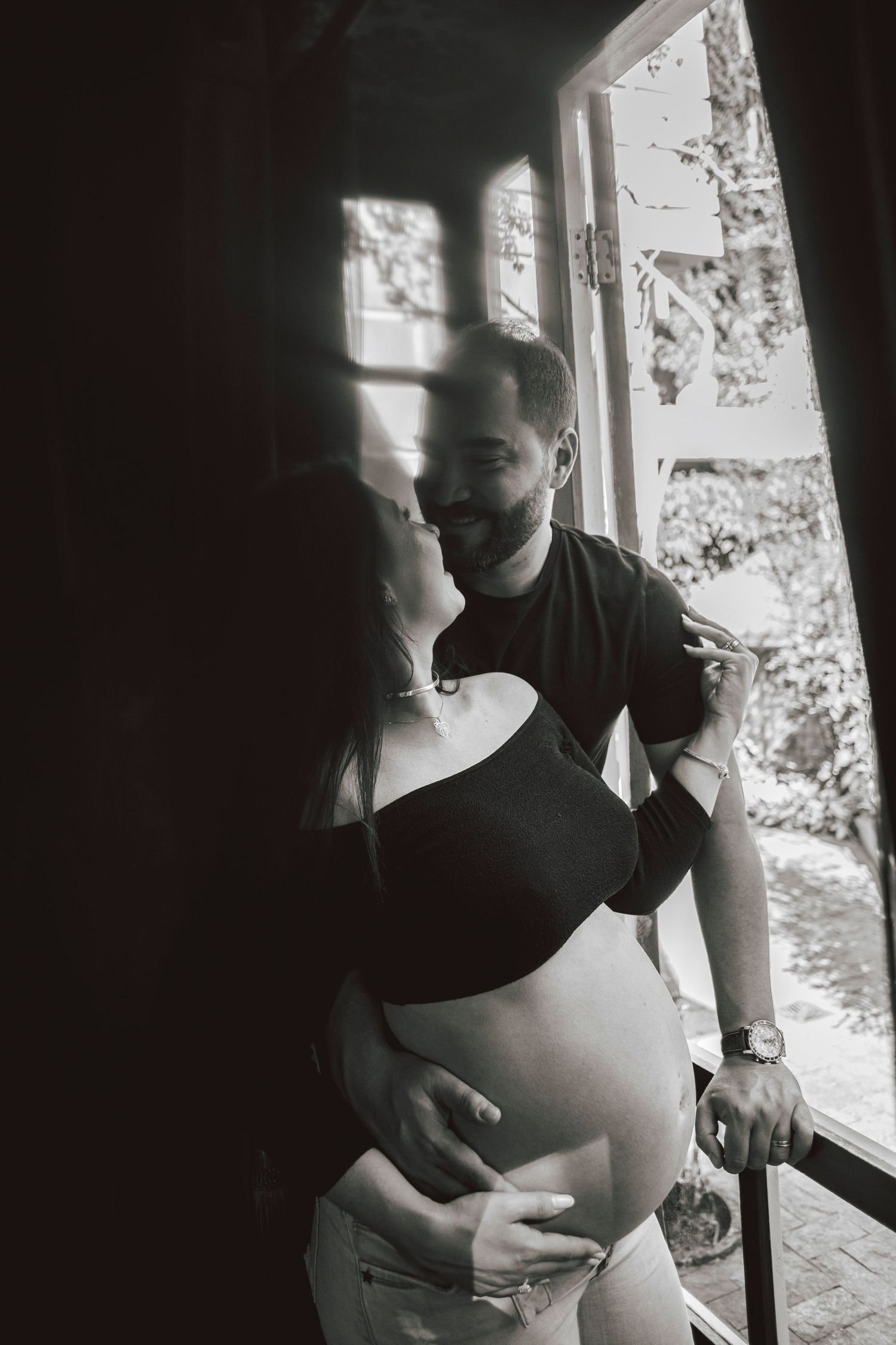 Intimate maternity photo of couple by window with natural light casting shadows.