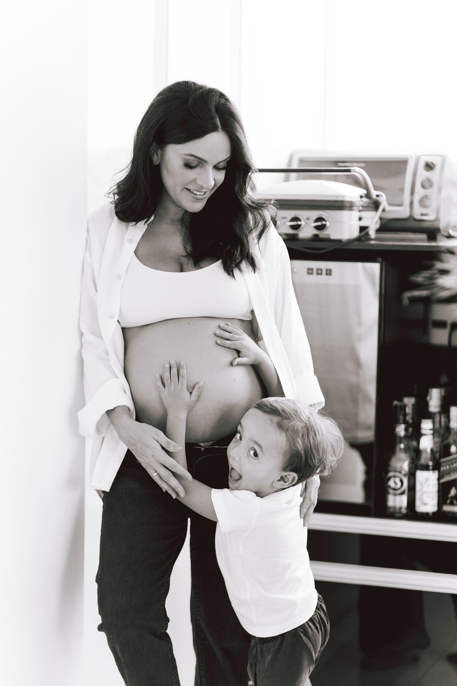 Heartwarming maternity photo of young child touching mother's pregnant belly in kitchen setting.