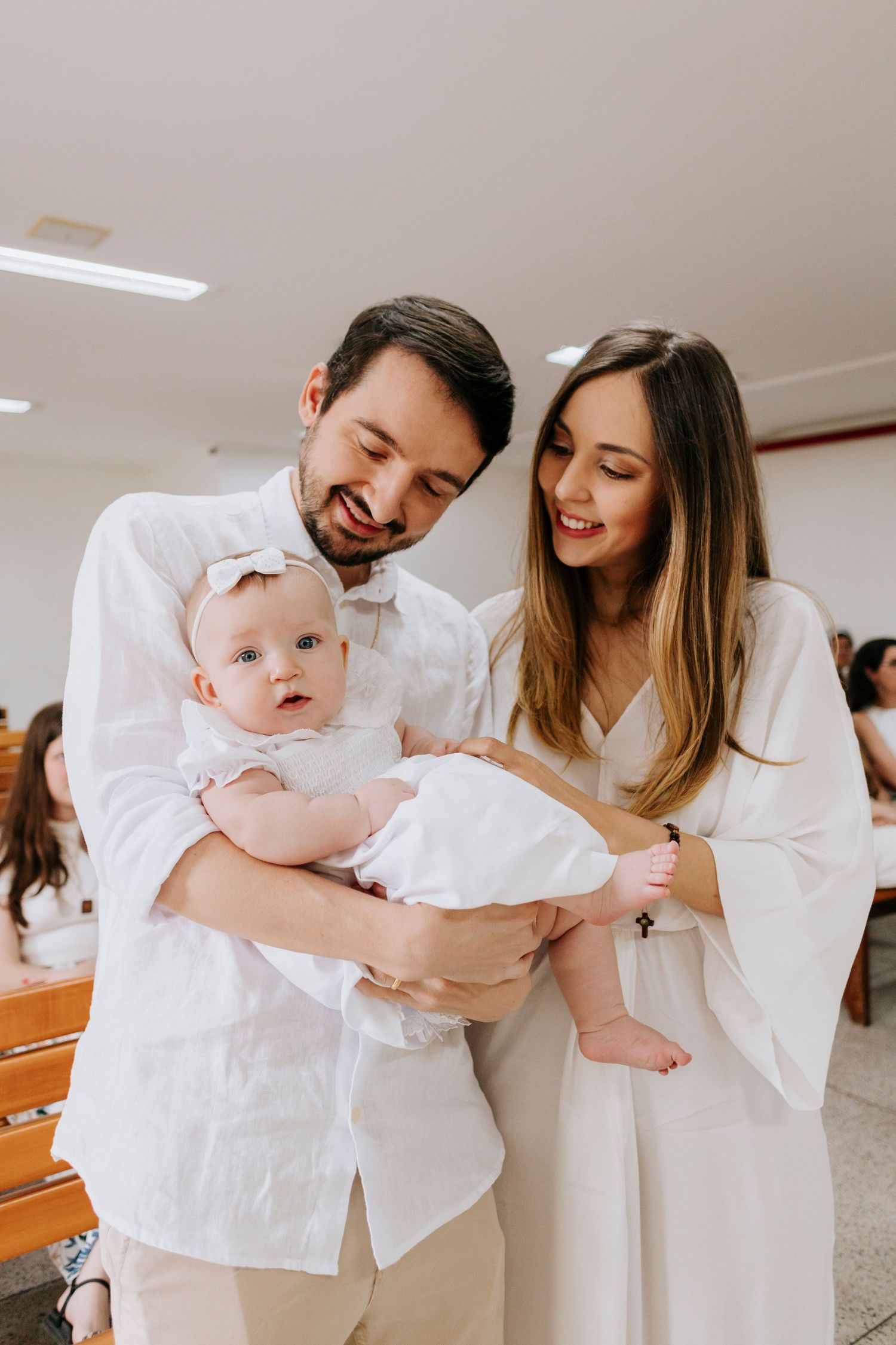 Parents in white clothing holding and smiling at their baby during special occasion.