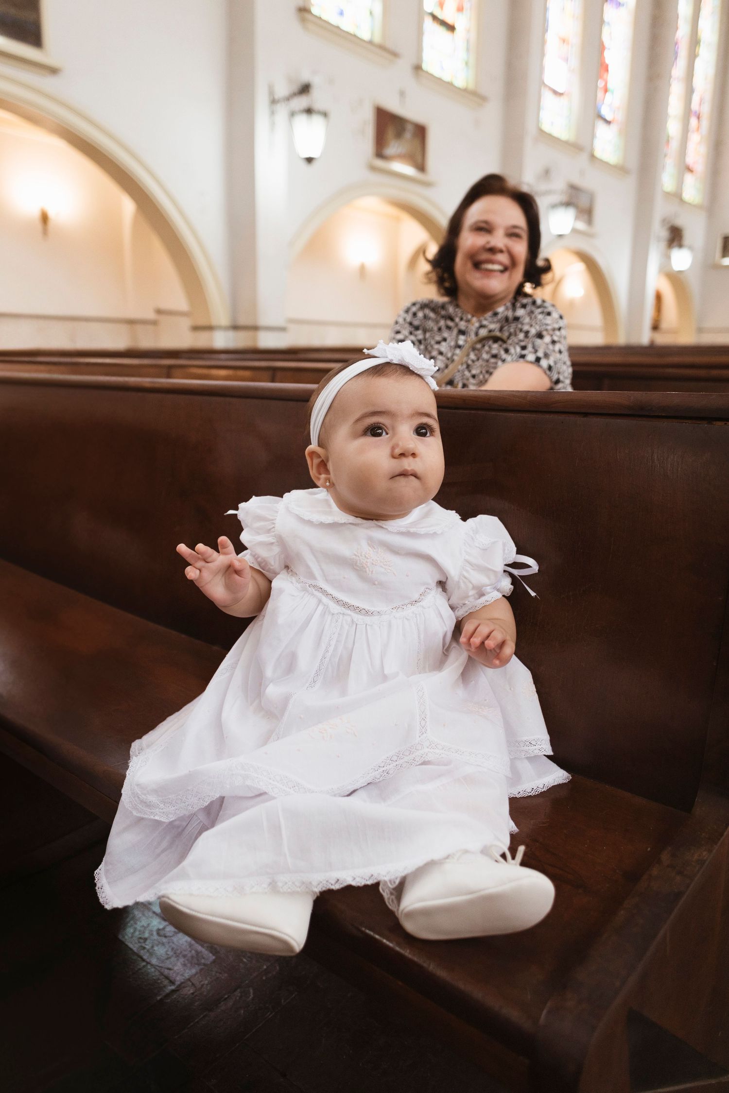 Baby in white christening dress sits on church pew during ceremony.